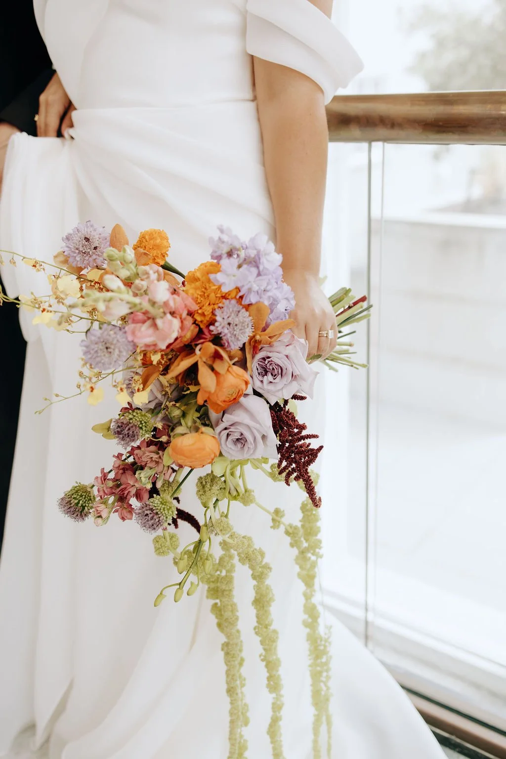 A bride in a white wedding dress holding a colorful bouquet of flowers, standing near a window with a wooden railing.