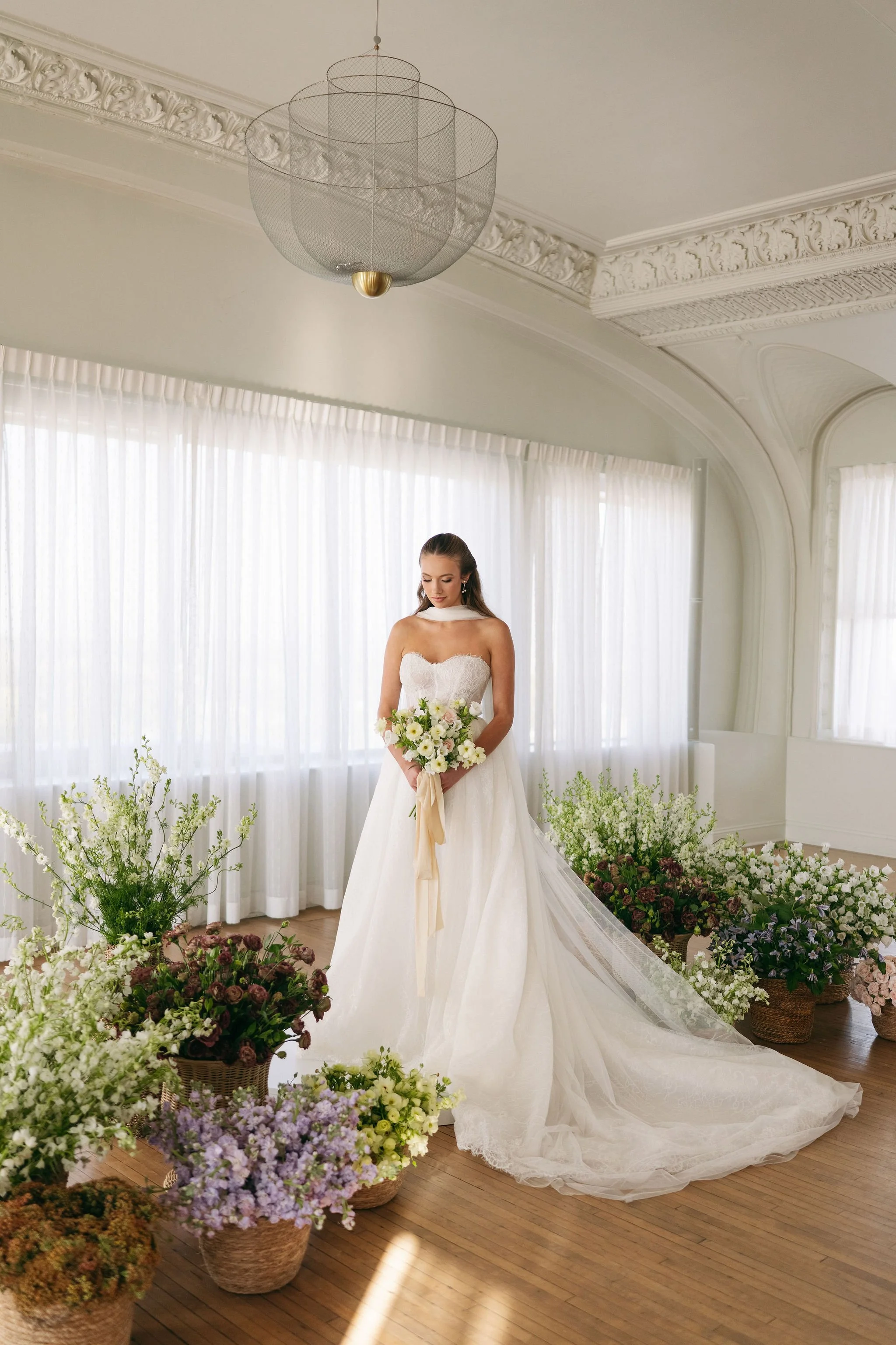 A bride in a white wedding dress stands in a bright room surrounded by an assortment of flowers.