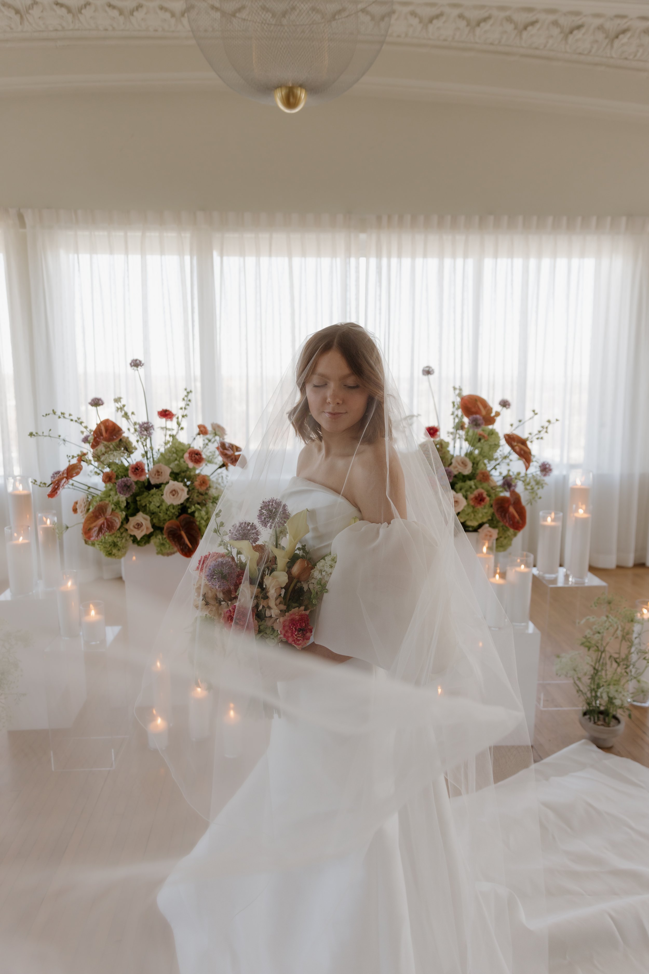 A bride in a white wedding dress with a veil holding a bouquet of flowers, standing in a room decorated with large flower arrangements and lit candles.