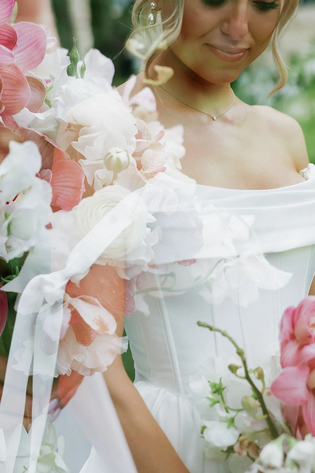 A woman in a white wedding dress holding a bouquet of pink and white flowers with greenery, smiling softly in an outdoor setting.