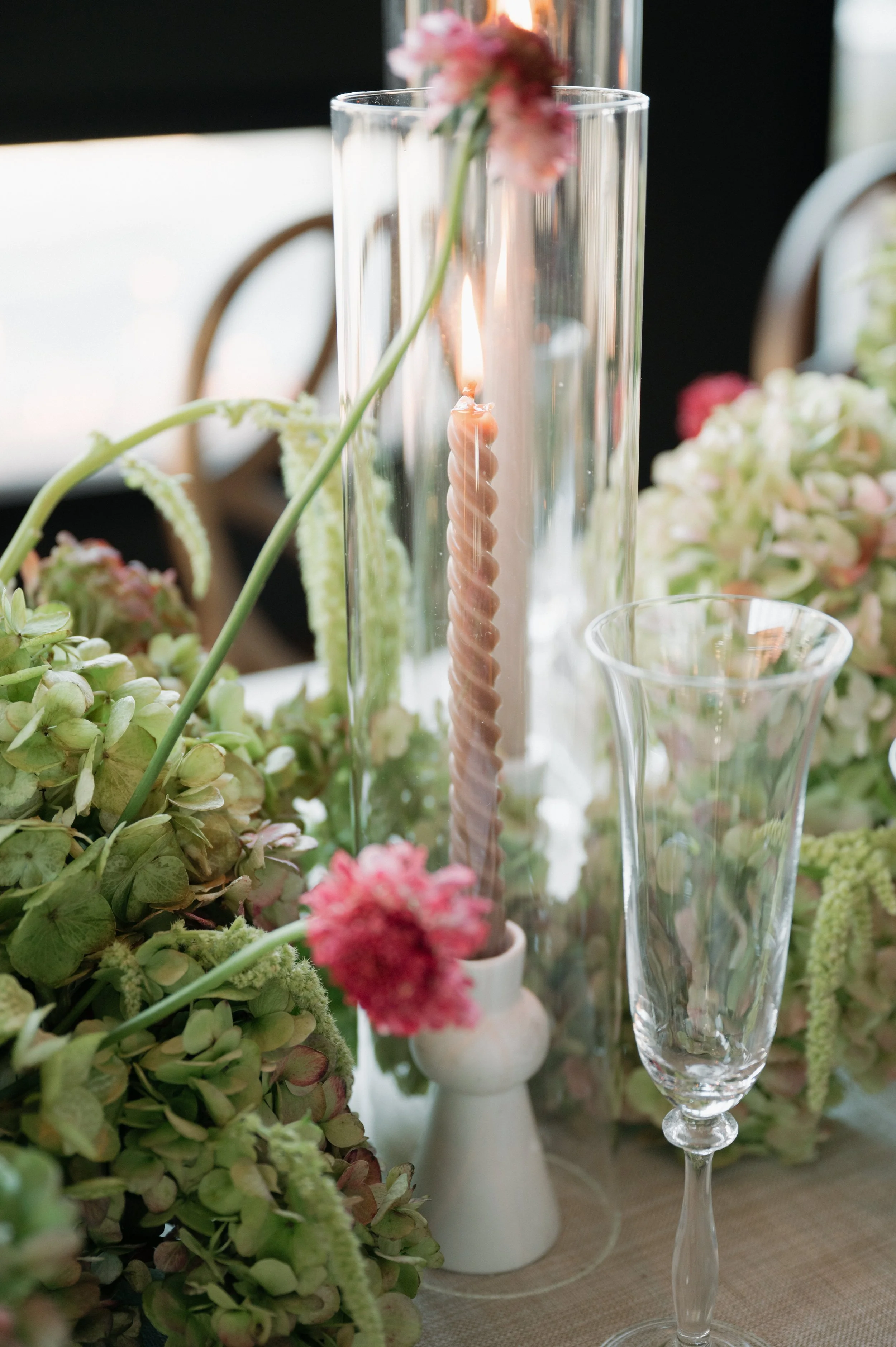 A table decorated with green hydrangeas, pink flowers, a tall glass candle holder with a lit pink twisted candle, and a clear champagne flute.