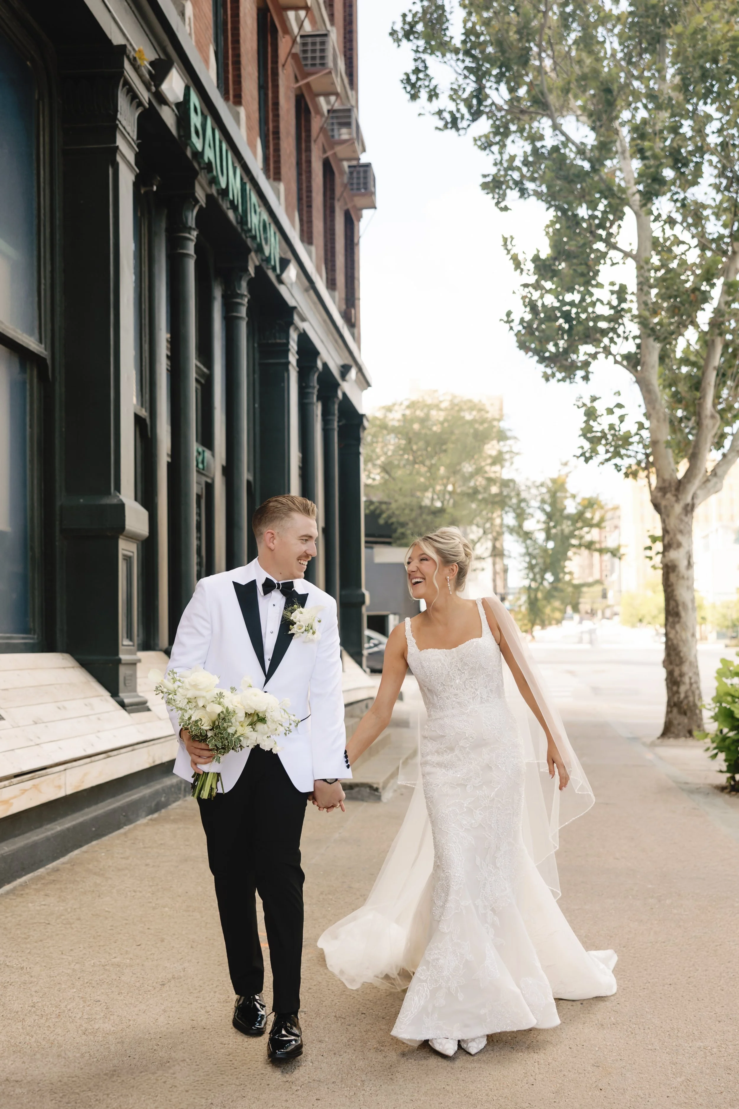 A newlywed couple walking hand in hand on a city sidewalk, smiling and laughing. The groom is wearing a white tuxedo jacket with black lapels, black pants, and holding a bouquet of white flowers. The bride is wearing a white lace wedding gown with a 