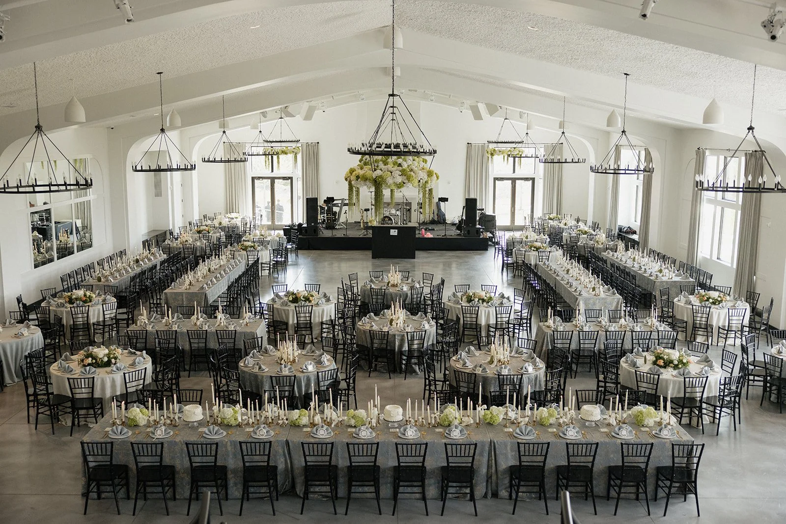 Wedding reception hall with round and rectangular tables decorated with white tablecloths, floral centerpieces, and candles, arranged in front of a stage with floral decorations and musical equipment, illuminated by large windows and hanging chandeli