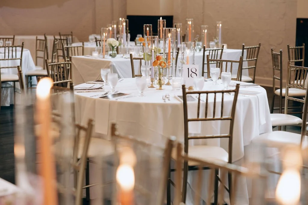 Elegant banquet hall decorated for a wedding reception with round tables covered in white tablecloths, gold chairs, tall candle holders, and floral centerpieces.