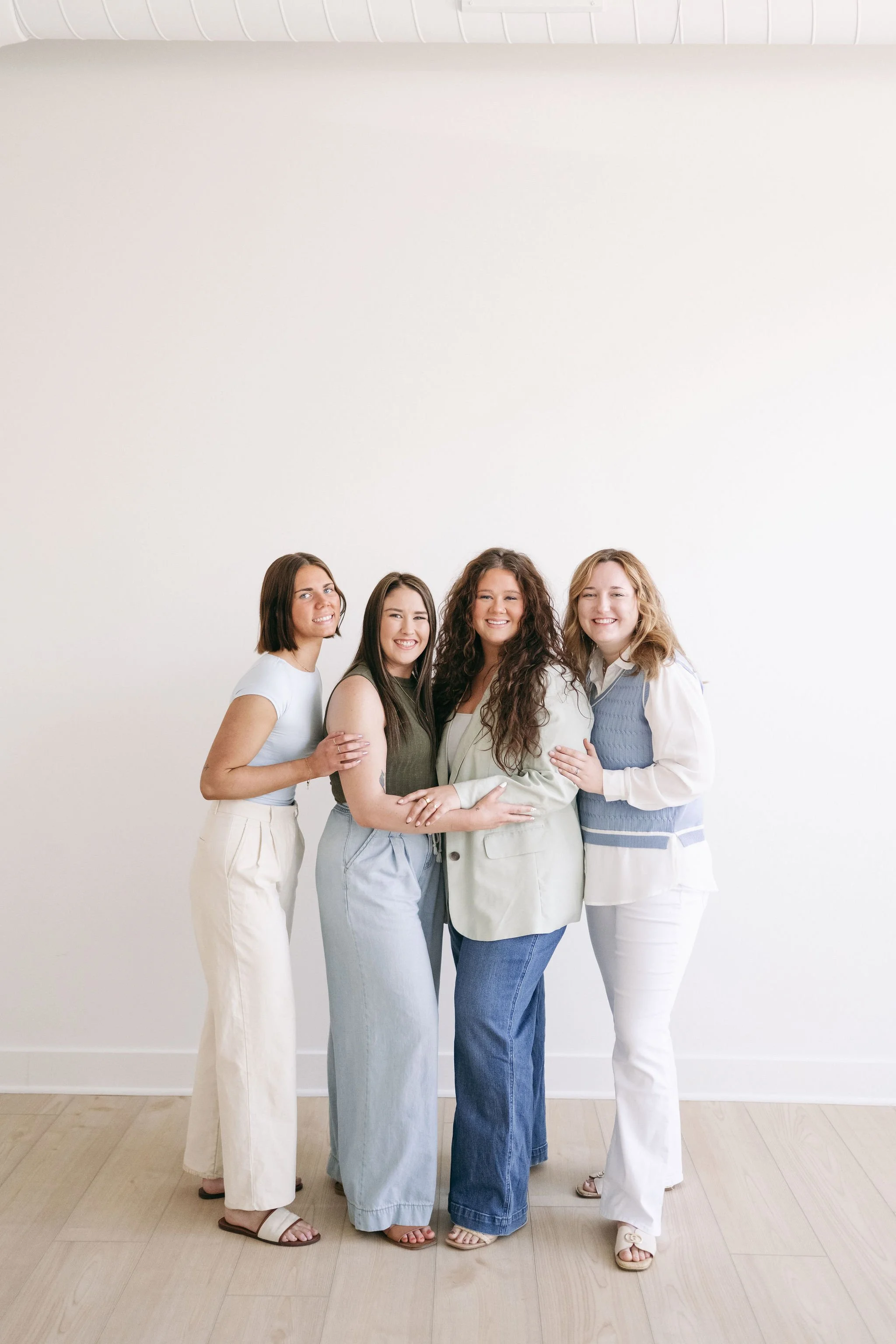 Four diverse women standing together against a white wall, smiling, with their arms around each other.