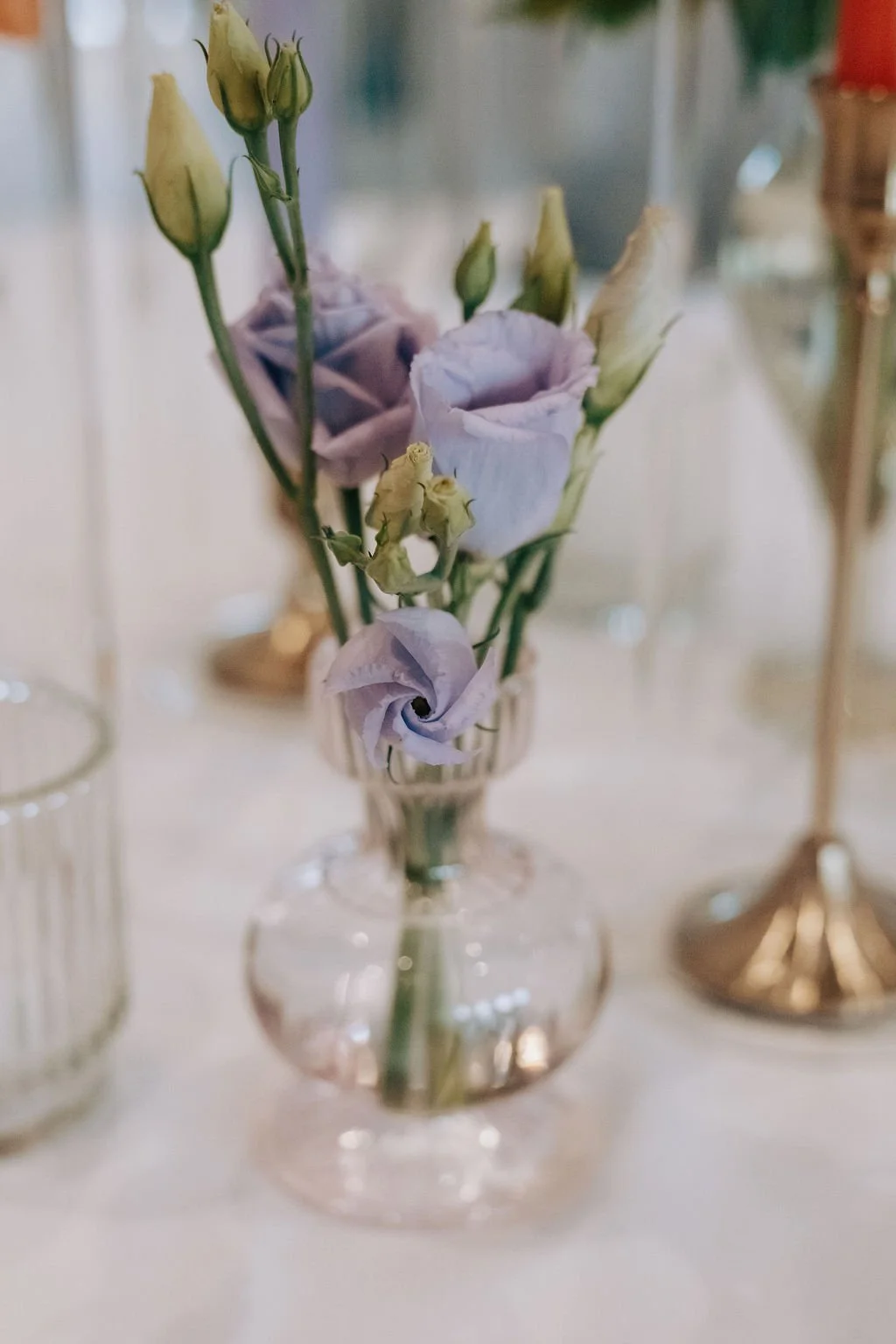A small glass vase holding a delicate arrangement of light purple flowers and buds, with a blurred background suggesting a decorative setting.