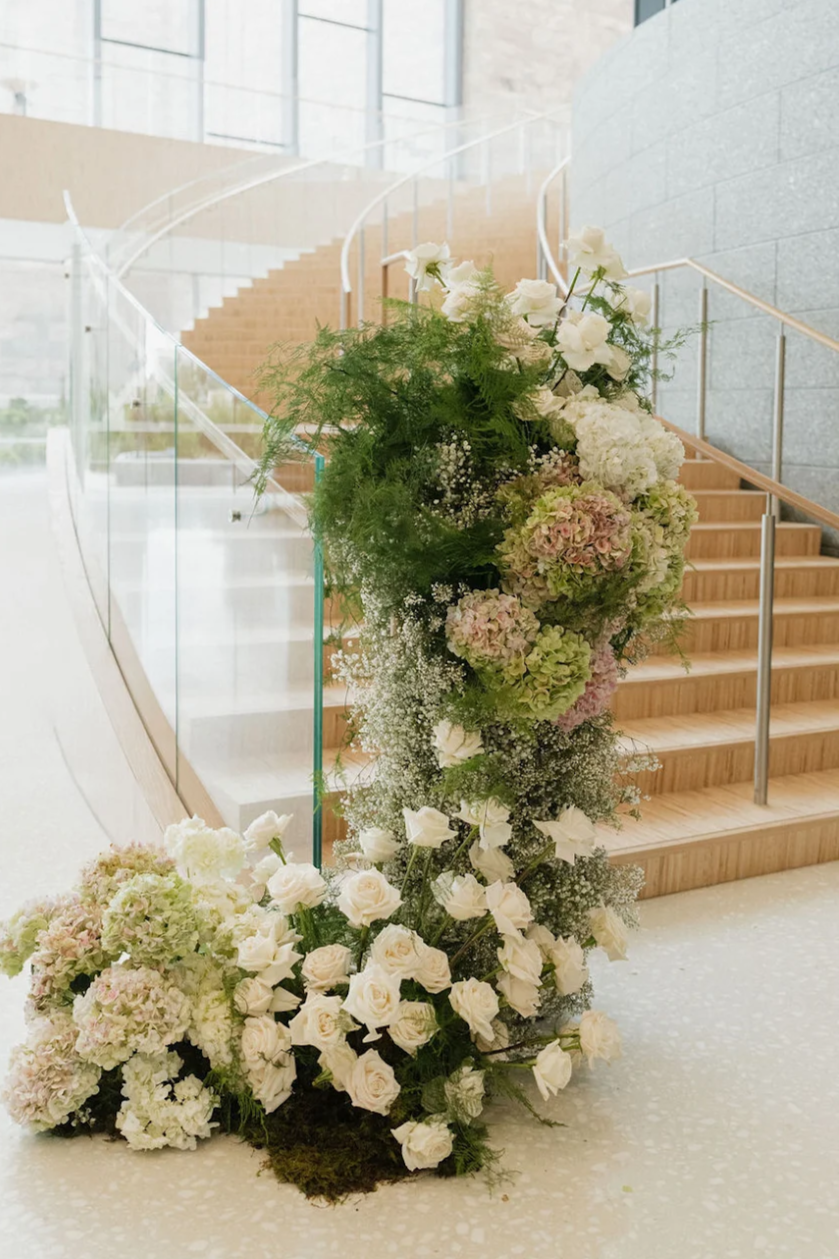 A floral arrangement with white and pastel pink flowers, greenery, and moss, placed at the base of stairs indoors.