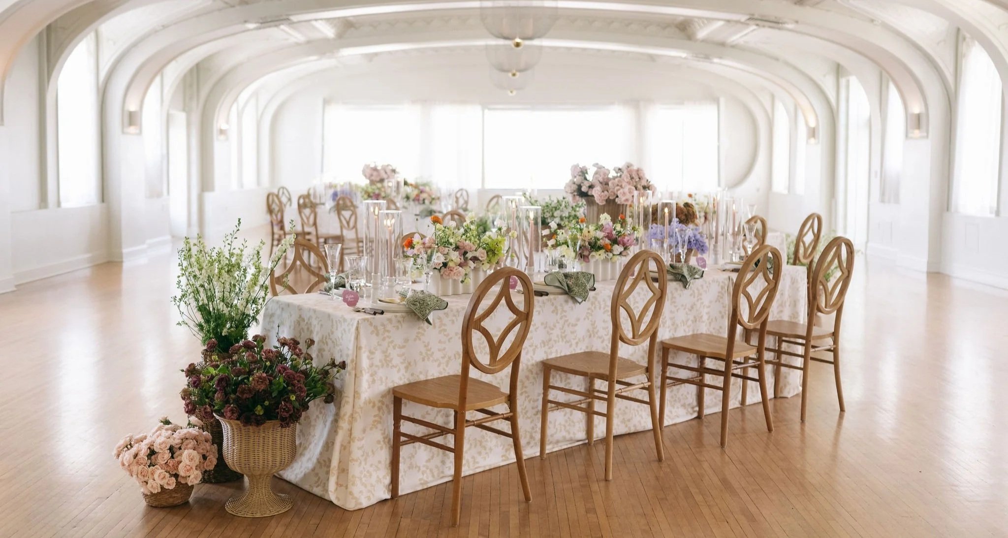 Elegant banquet table set with floral centerpieces and candles in a bright, spacious room with large windows and arched ceilings.