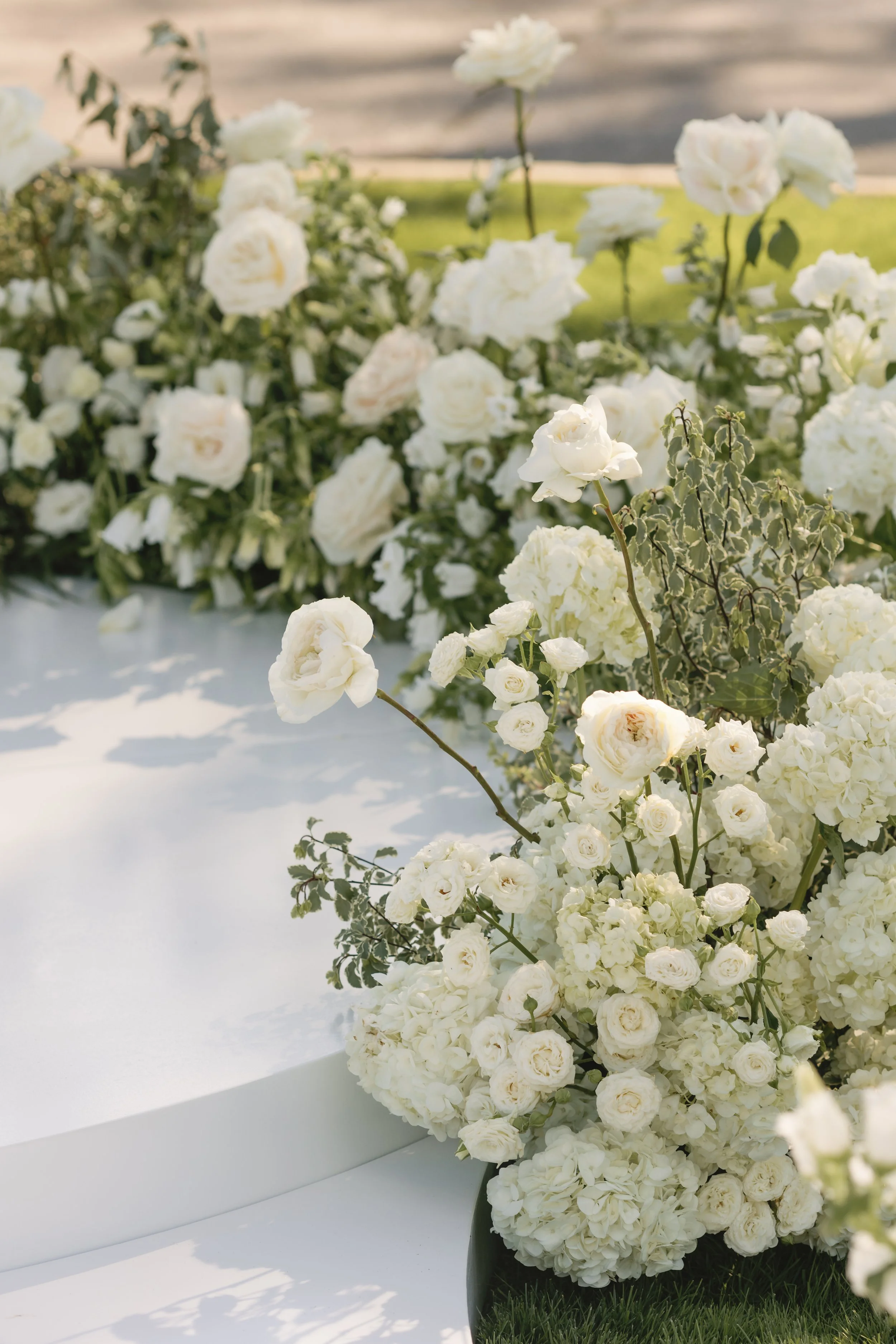 White floral arrangement with roses, hydrangeas, and other flowers at an outdoor event with a green lawn and a paved walkway in the background.