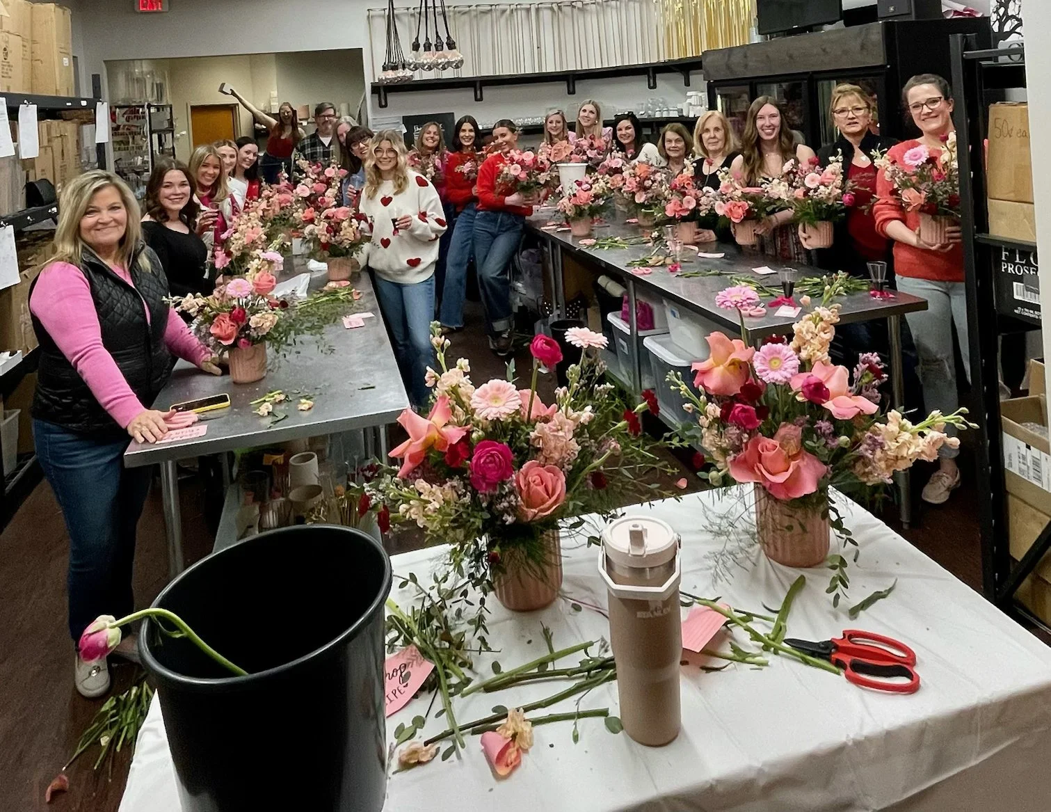 Group of women gathered in a flower arranging workshop, holding pink and peach flower arrangements, with floral supplies and scissors on the table, and shelves of boxes in the background.