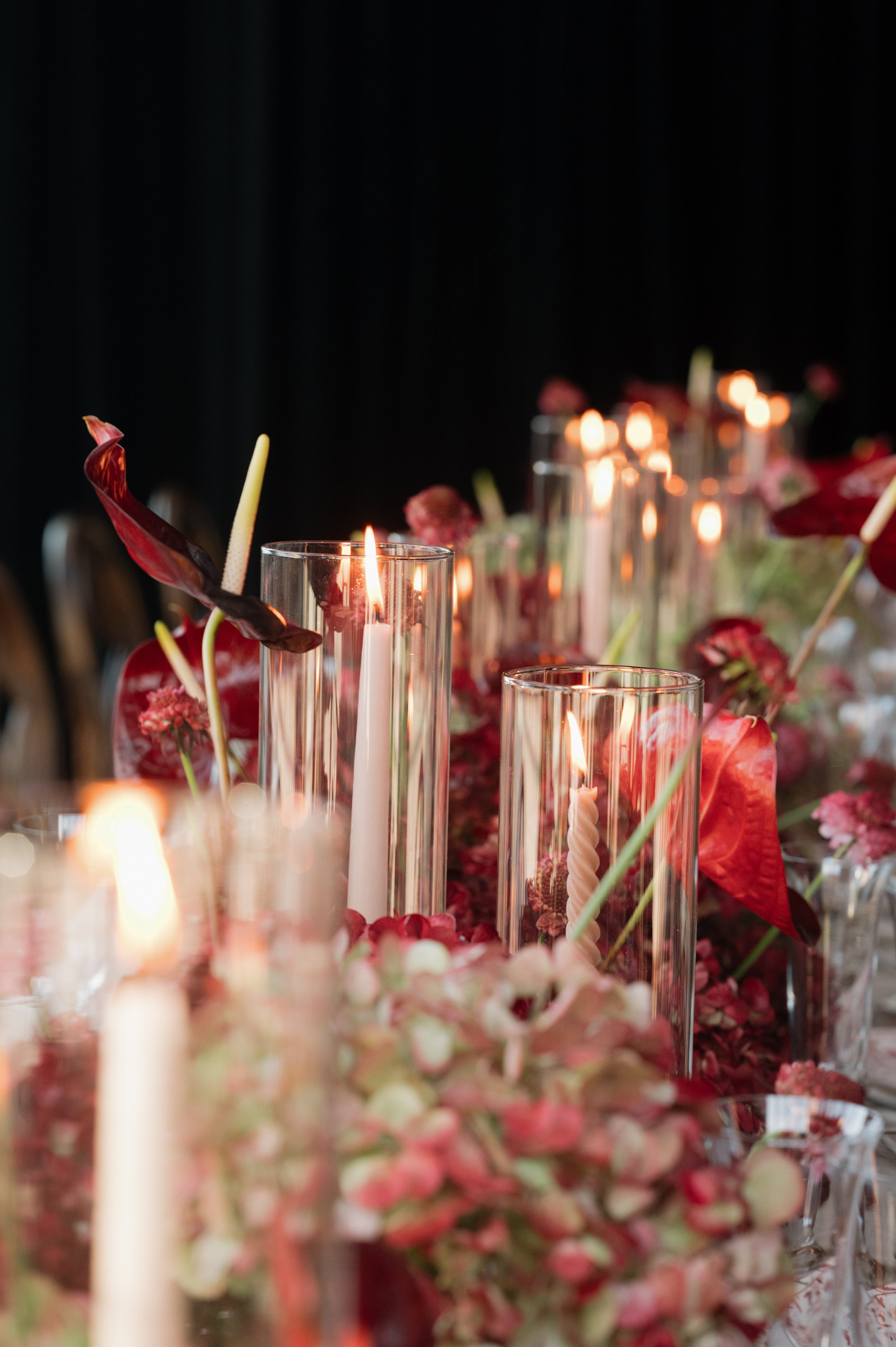 A close-up of a decorated table with tall glass candle holders, lit candles, and floral arrangements, featuring pink and red flowers with dark green foliage, set against a dark background.