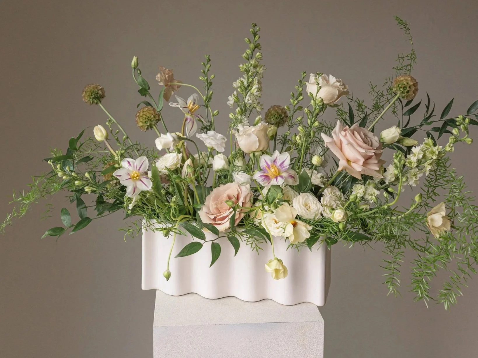 A floral arrangement in a white, scalloped ceramic vase on a pedestal, featuring light pink roses, white roses, and various greenery and smaller flowers.