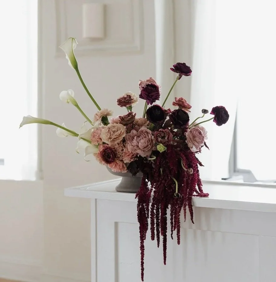 A vase of mixed flowers, including white calla lilies and dark purple and pink blooms, placed on a white surface near a window with natural light.