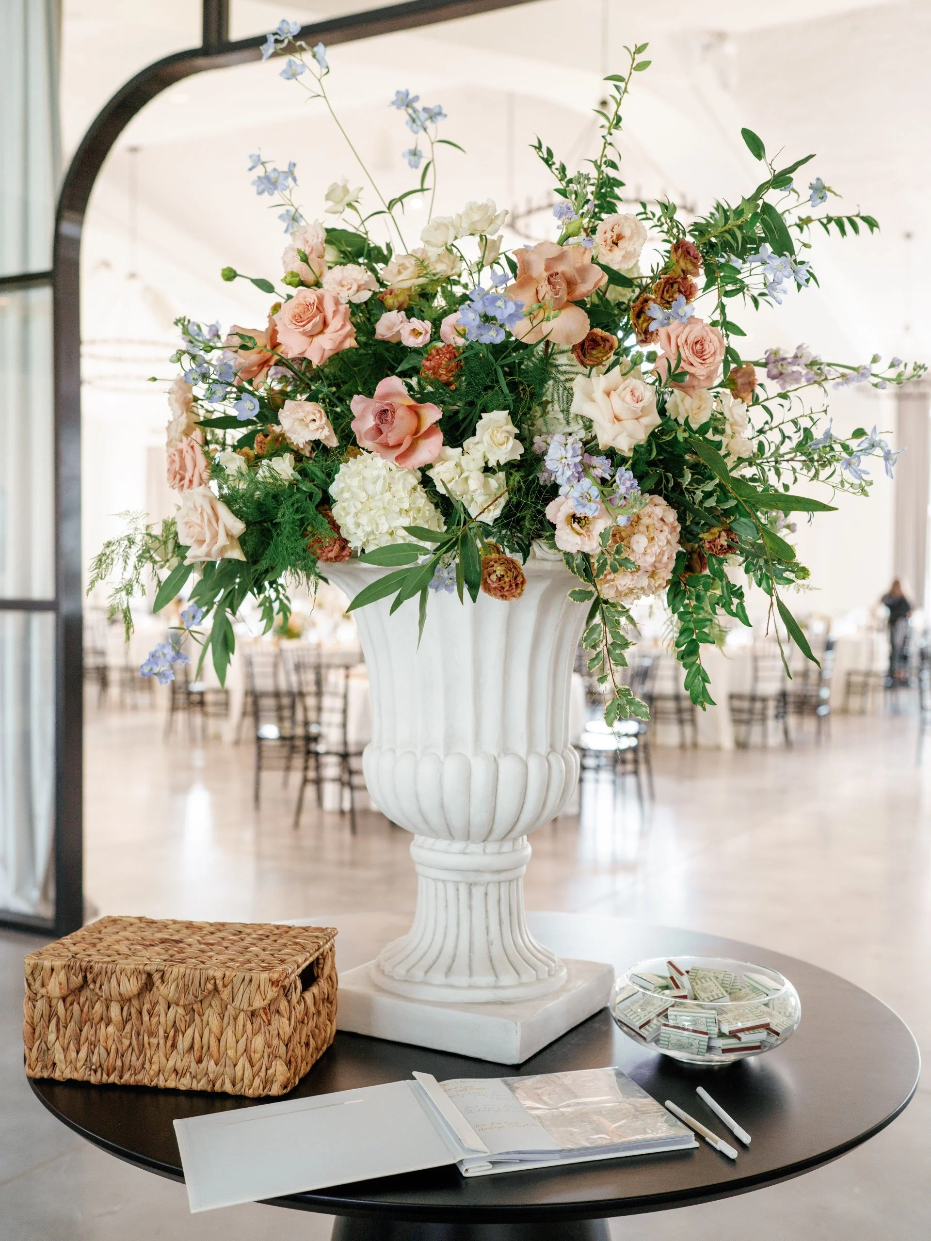 A large floral arrangement in a white urn with pink, white, and purple flowers, including roses and delphiniums, on a black table with a woven basket, a guestbook, and two pens, in a bright event space with tables and chairs in the background.