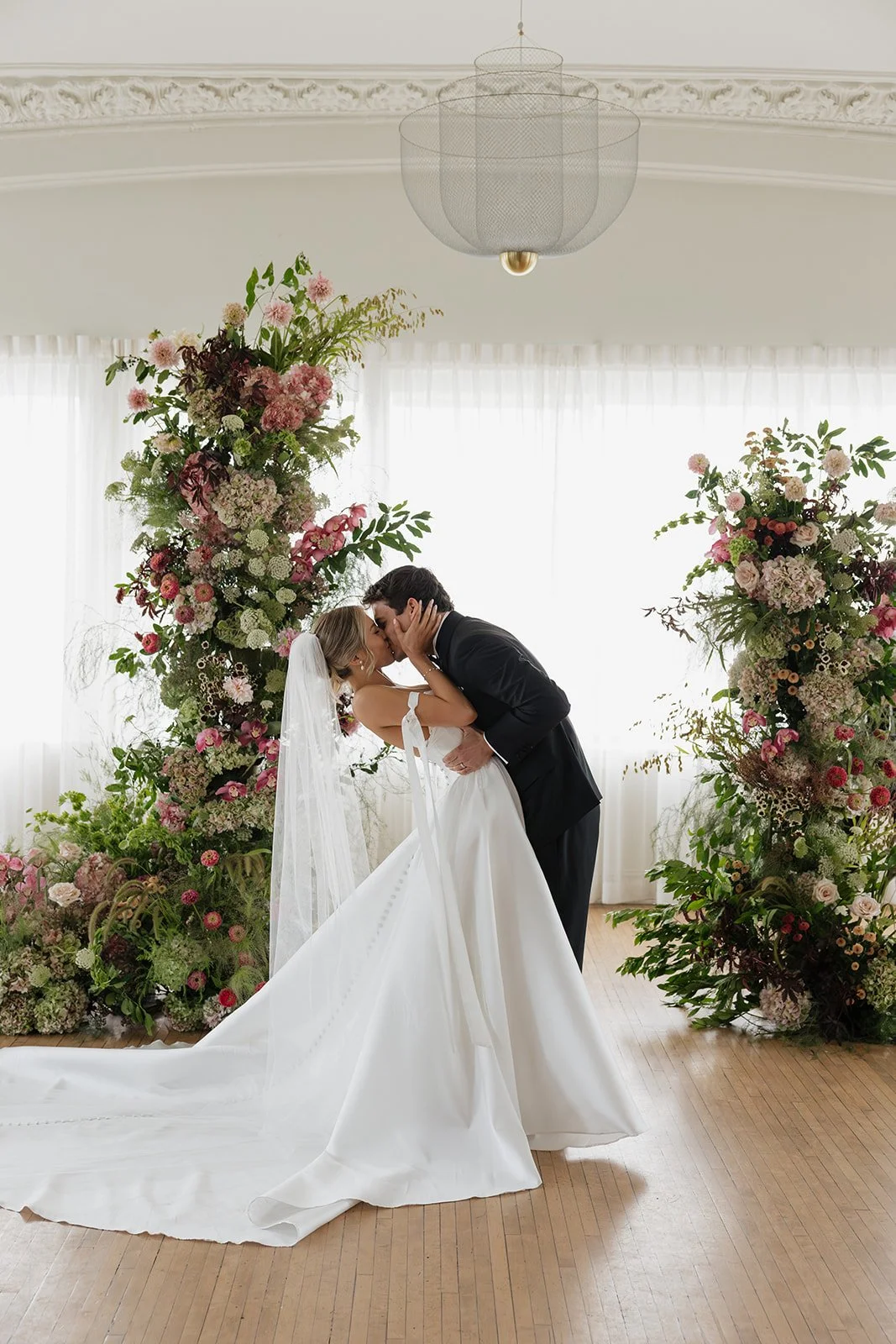 A bride and groom sharing a kiss during their wedding ceremony, surrounded by tall floral arrangements with pink and white flowers, in a bright room with sheer curtains.