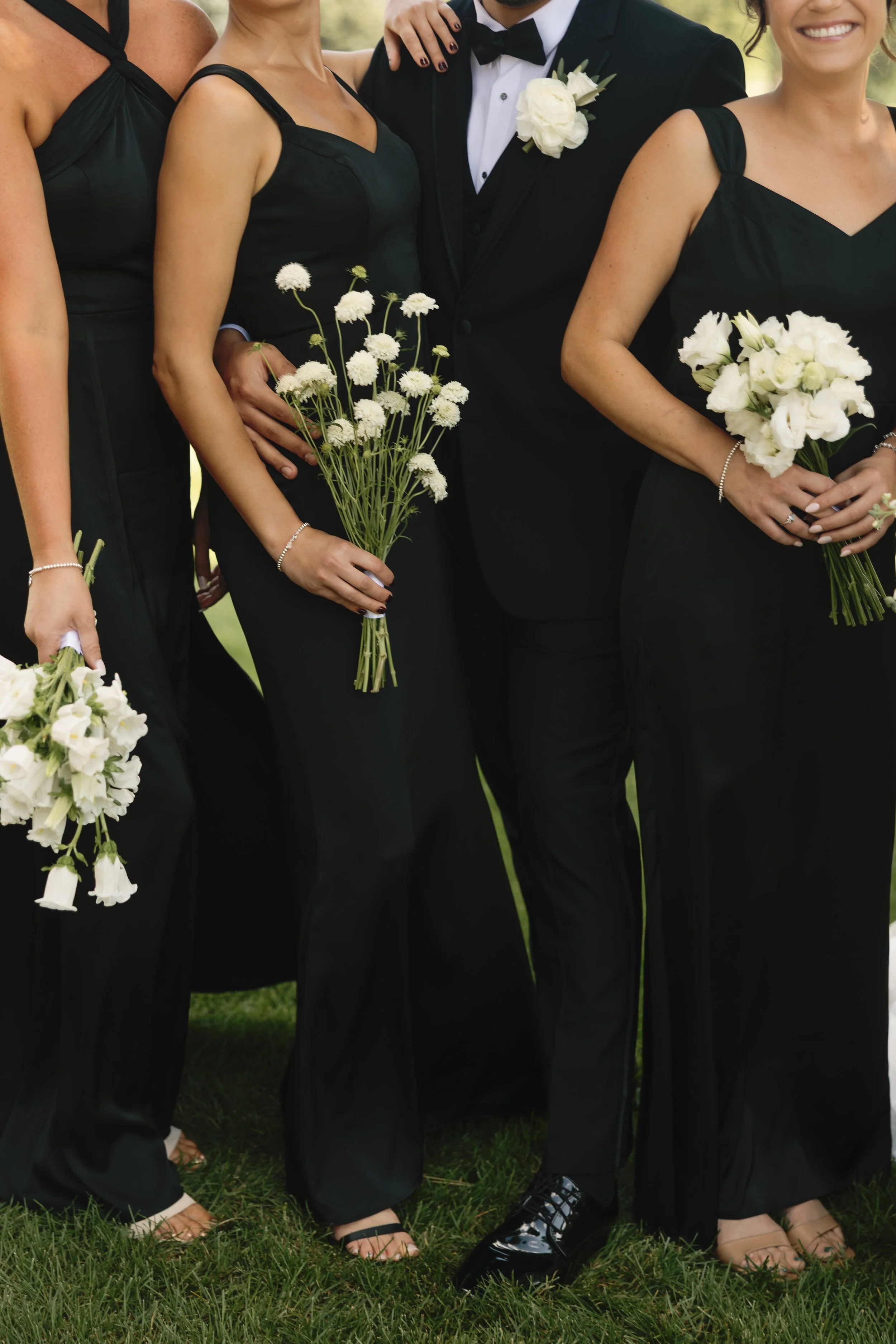 Close-up of a group of people dressed in formal black attire, holding white flower bouquets, standing outdoors on grass, likely at a wedding or formal event.