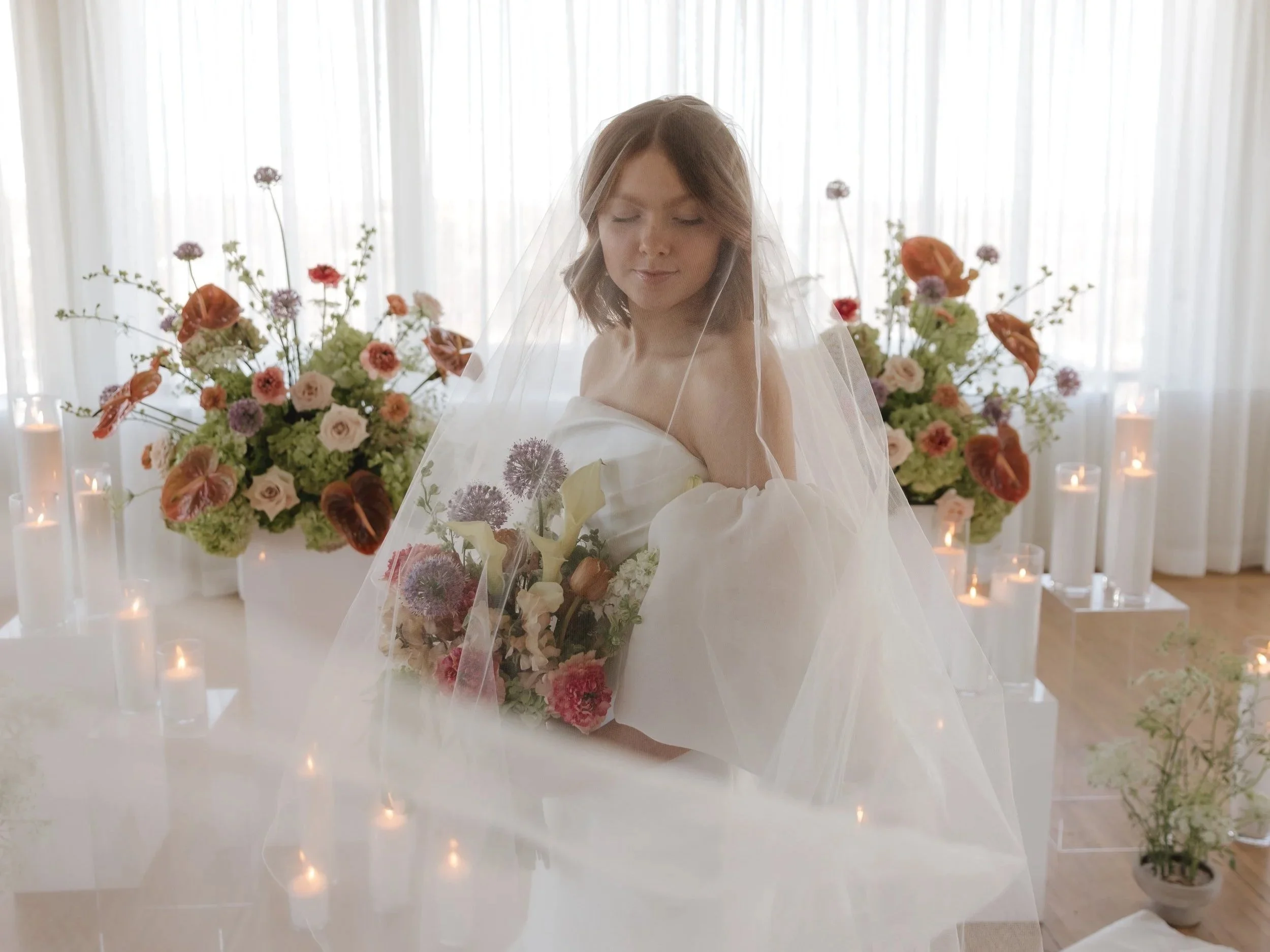 A bride in a wedding dress with a veil holding a bouquet of flowers, standing in front of large floral arrangements and candles.