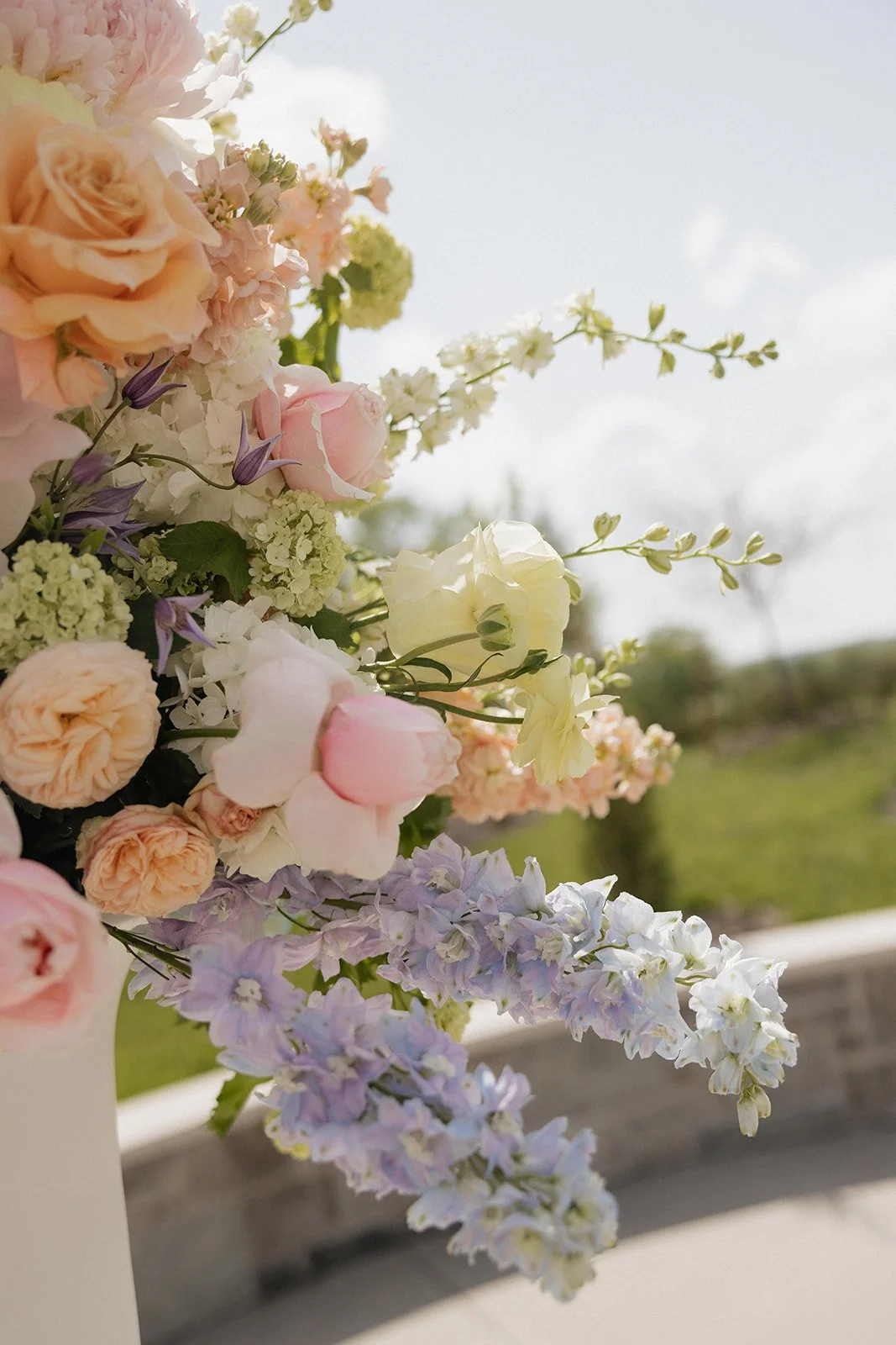 Close-up of a pastel-colored flower arrangement with roses, hydrangeas, and delphiniums outdoors, with a blurred background of green trees and blue sky.