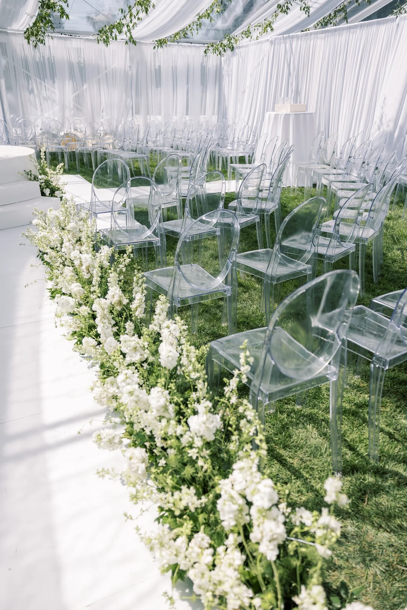 An outdoor wedding setup with clear acrylic chairs lined on green grass, white floral arrangements along a white aisle, and white draped fabric decorating the tent ceiling.