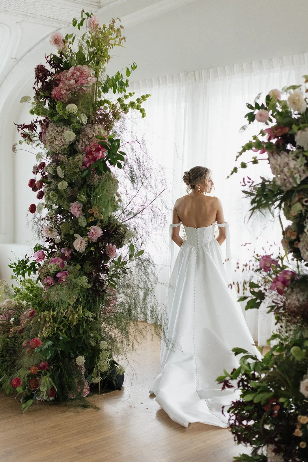 Bride in a white wedding gown standing beside a floral arch.