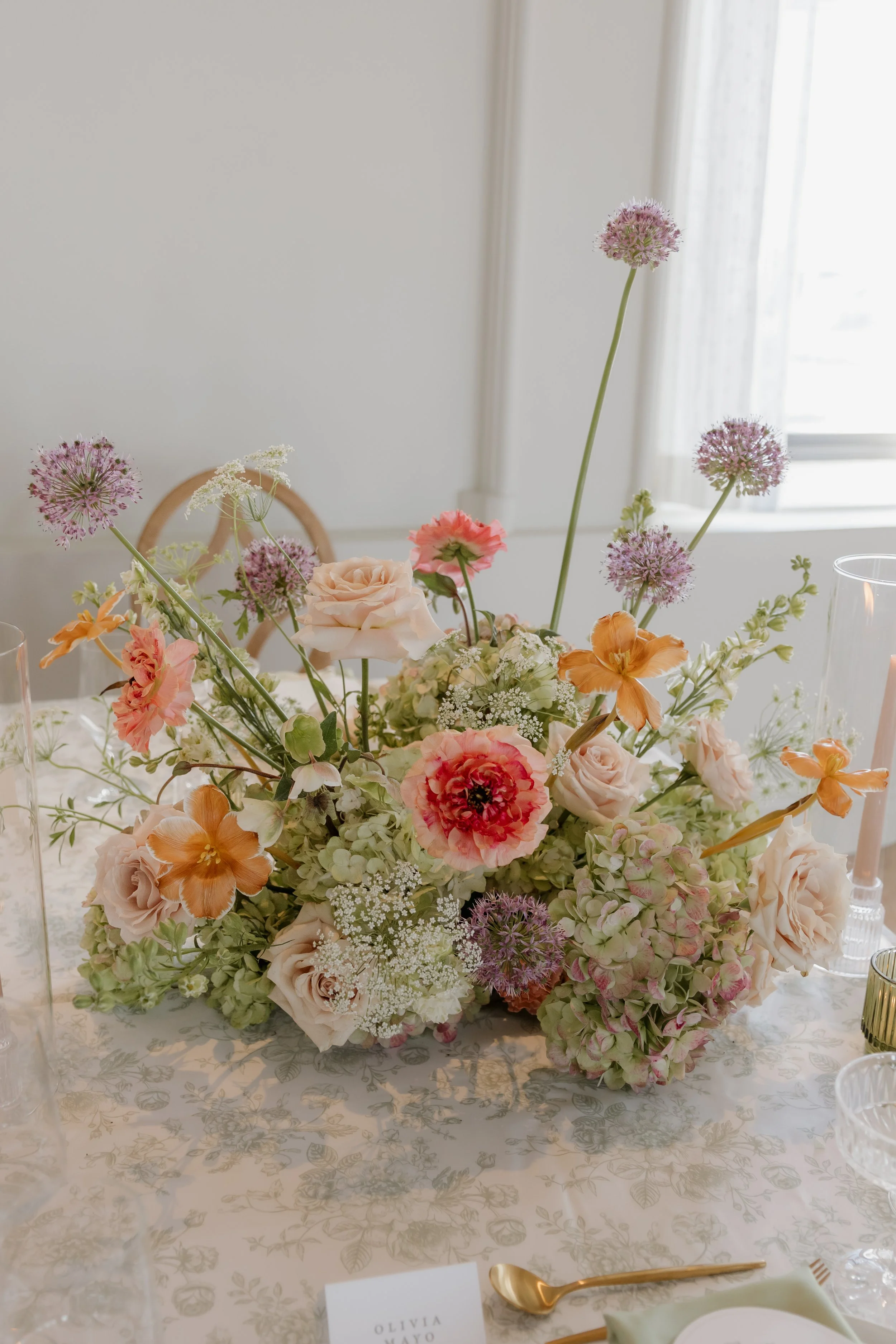 A floral centerpiece with pink, white, and peach flowers on a decorated table with gold utensils and glassware.