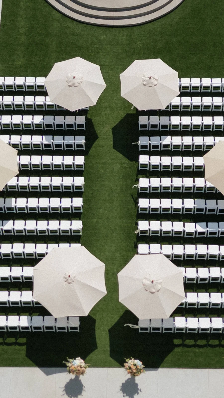 Arrangement of white chairs and large white umbrellas on a grassy outdoor area with landscaped flower arrangements at the bottom.