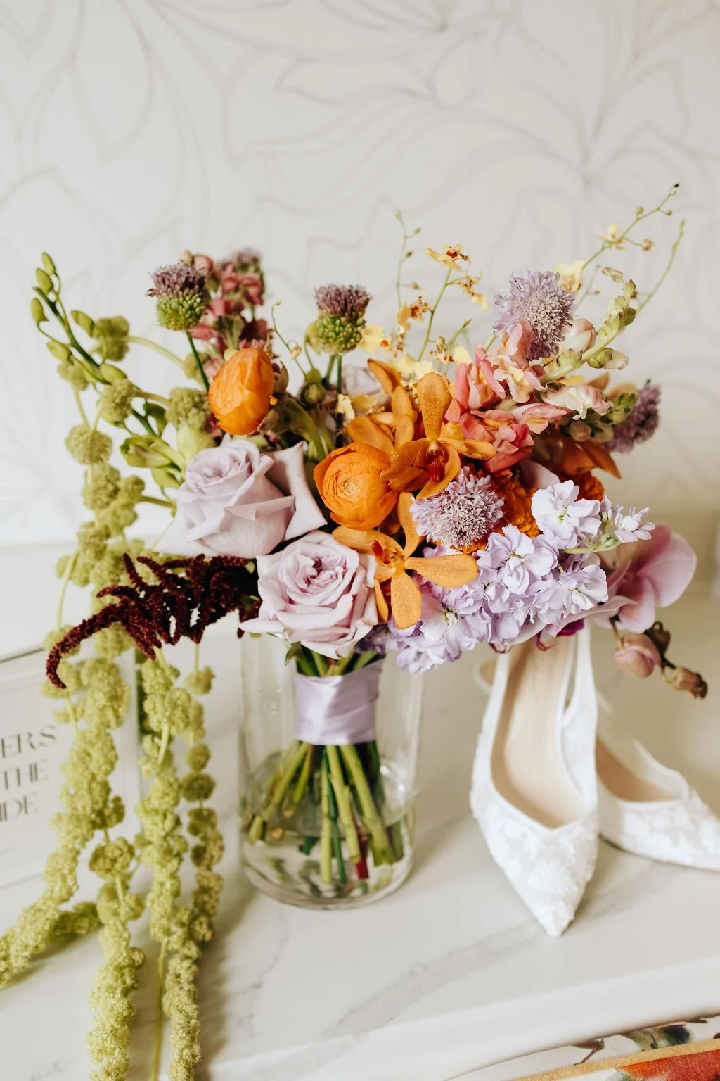 A colorful bouquet of mixed flowers placed in a clear glass vase, with a pair of white lace high heel shoes next to it.