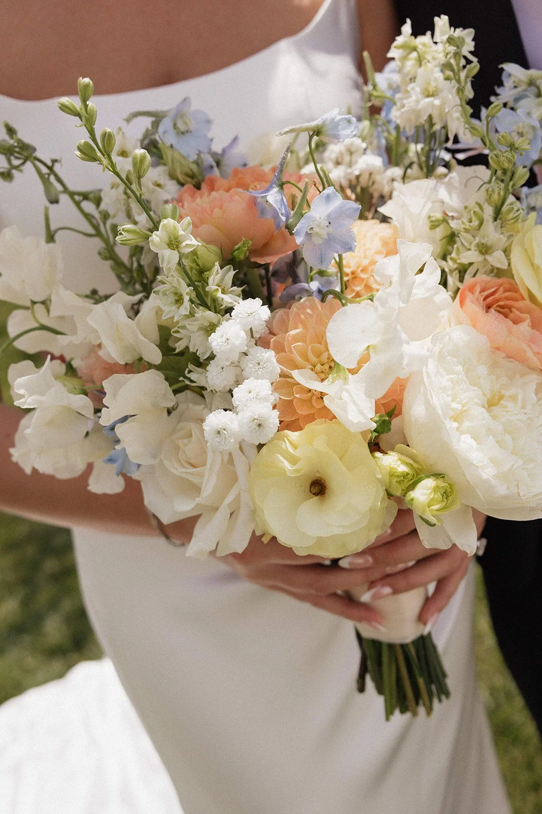 Close-up of a bride holding a bouquet of pastel-colored flowers, including white, peach, and light blue blossoms, at a wedding.