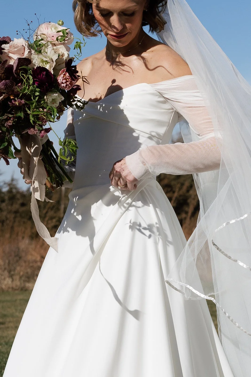 A bride in a white wedding dress and veil holding a bouquet of flowers outdoors on a sunny day.