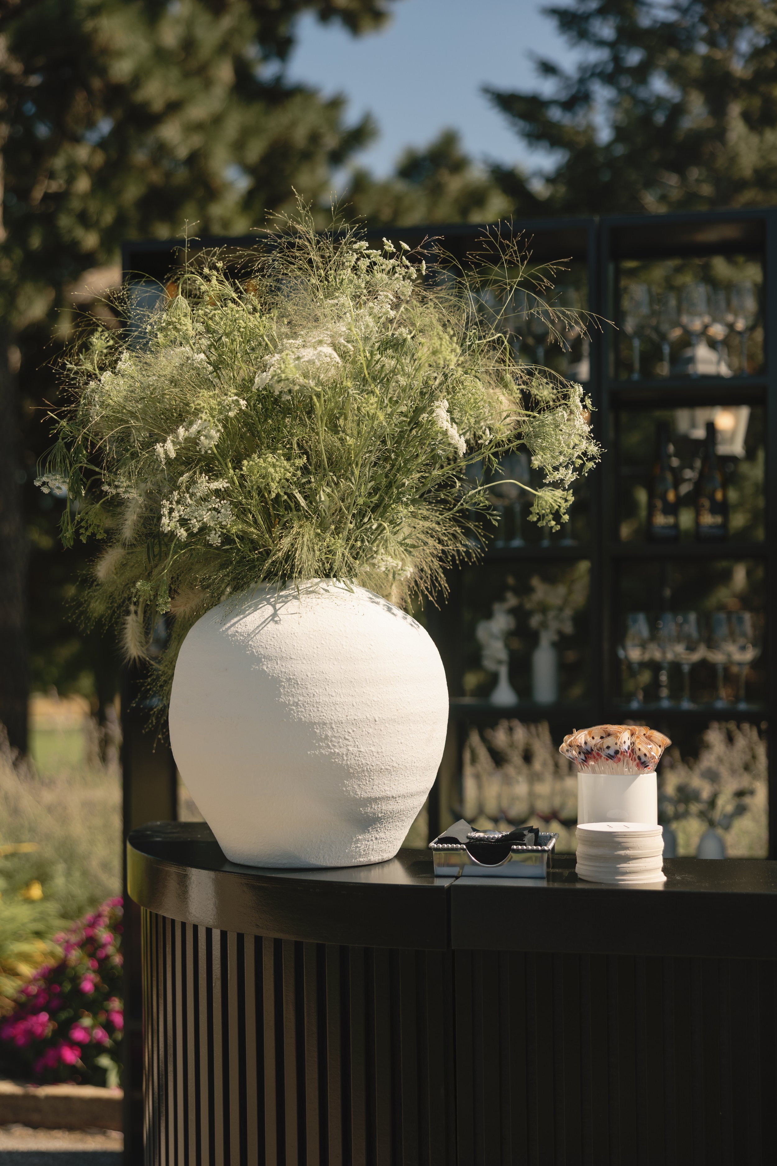 Large white vase filled with various green and white wildflowers on a black table outdoors.