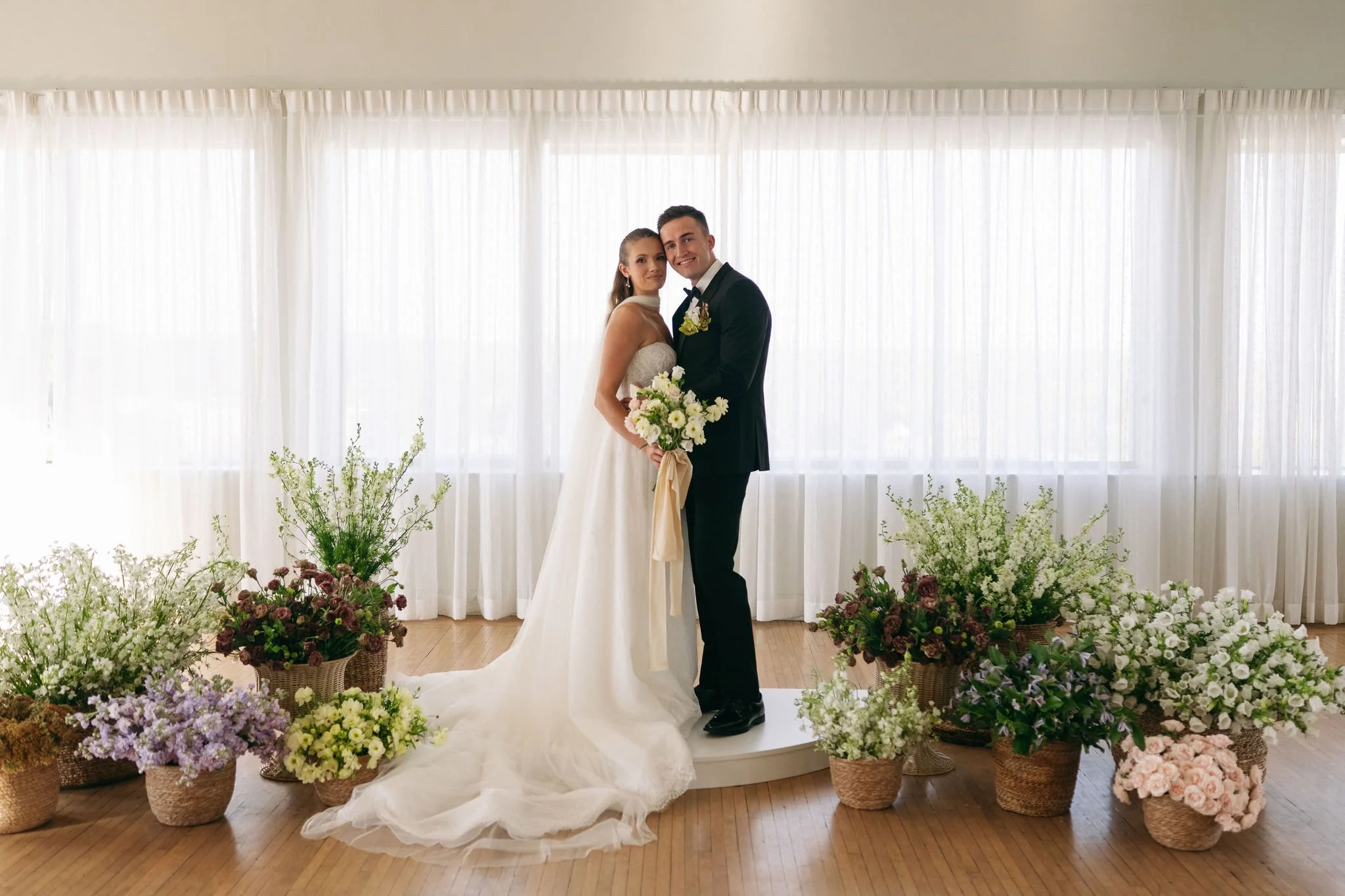 A bride and groom standing together on a white platform, surrounded by an arrangement of flowers in baskets, with white curtains in the background.