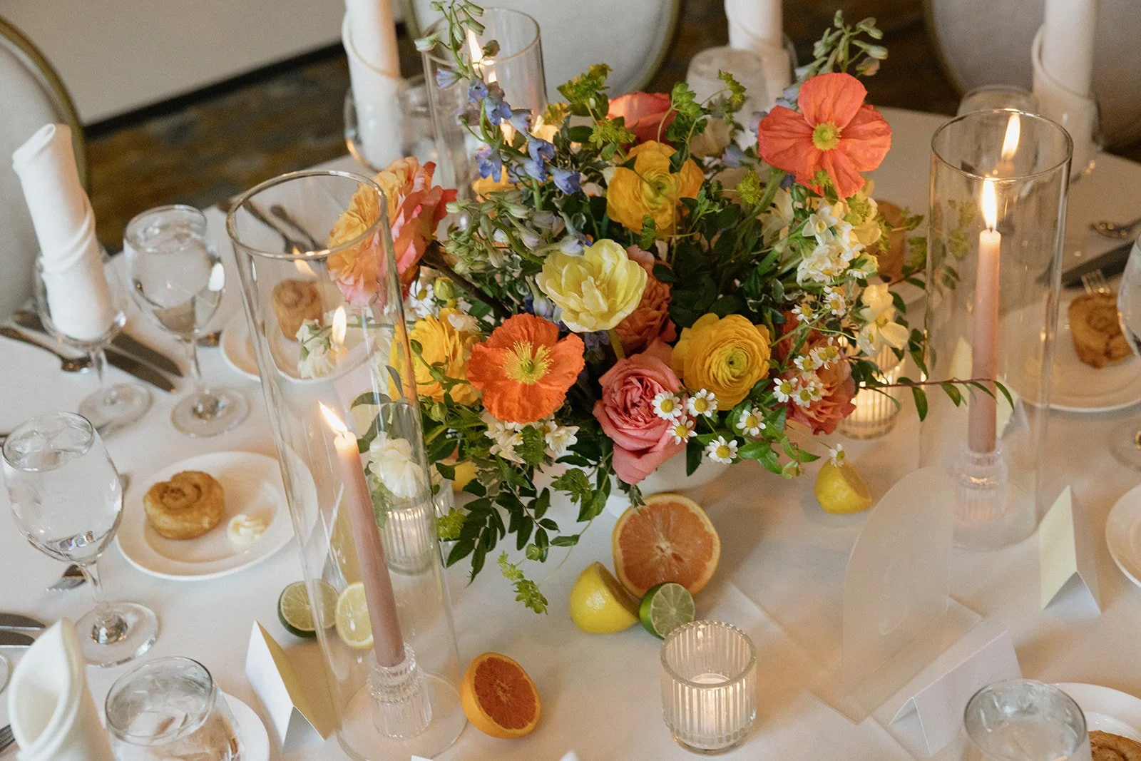A wedding or event table centerpiece with colorful flowers including roses, poppies, and daisies, surrounded by citrus fruits, candles, and glassware.