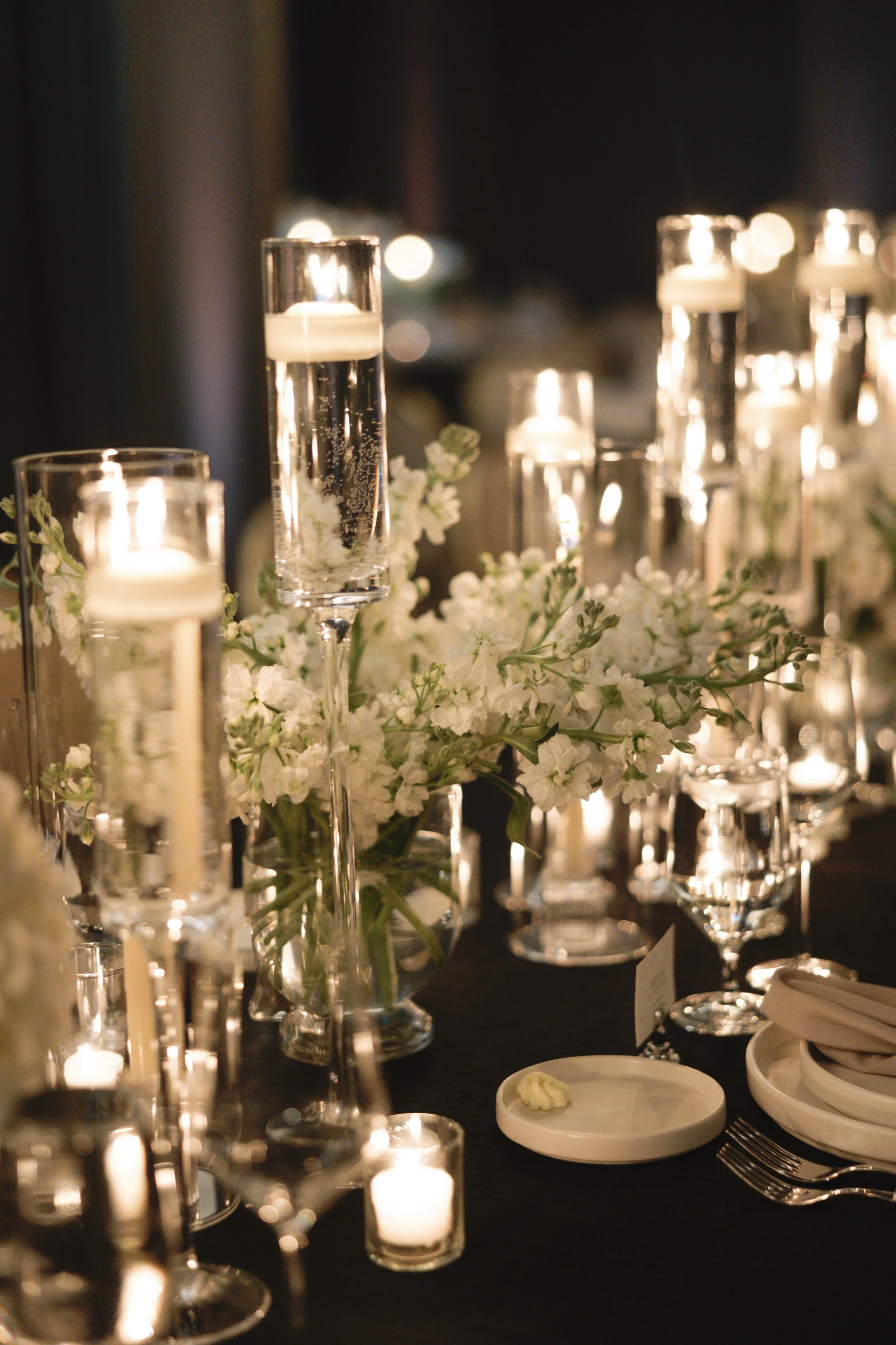Elegant table setting with lit candles in glass holders, white flowers, and dinnerware for a formal event.
