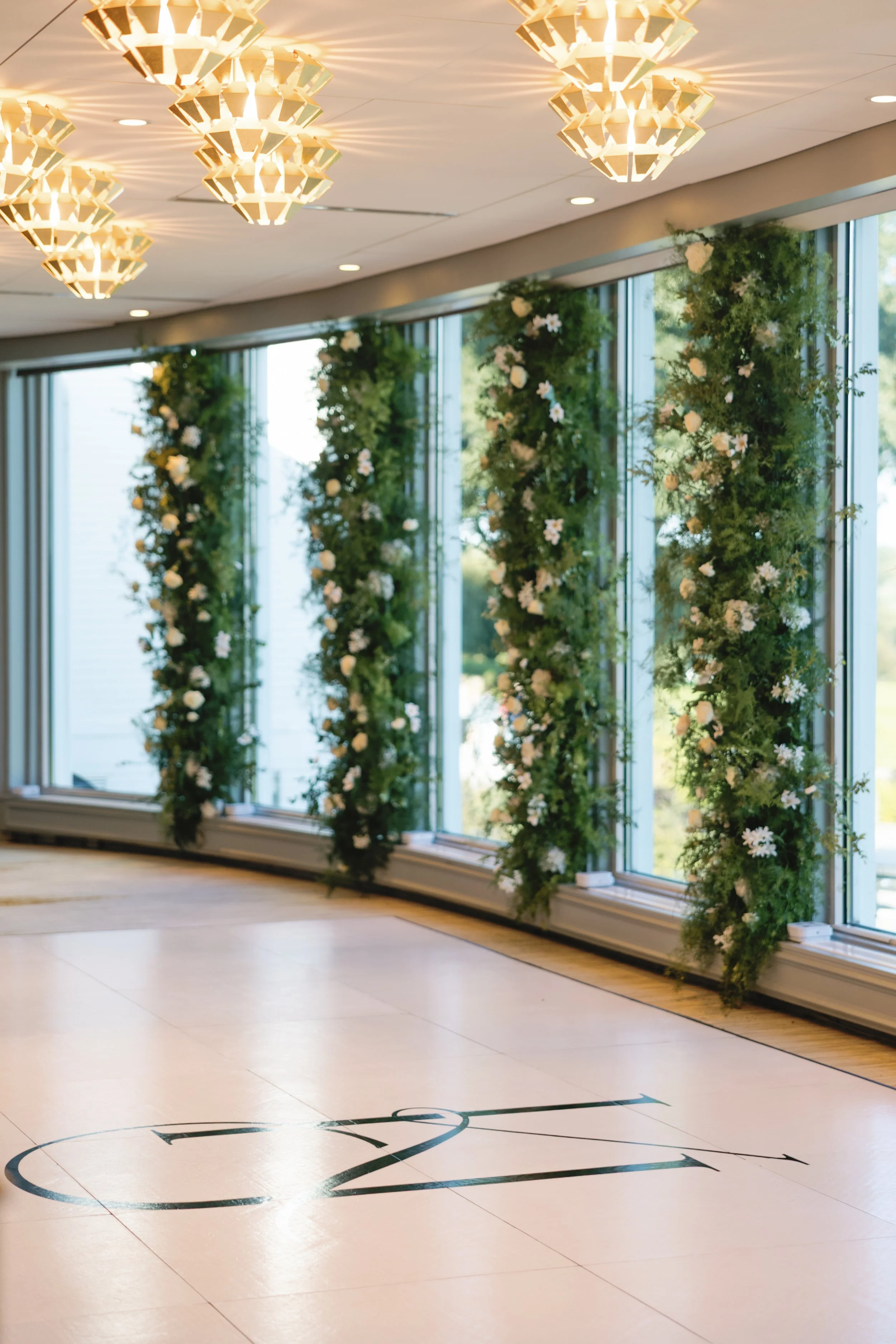 Indoor event space decorated with greenery and white flowers, windows in the background, and a black dance floor with a large 'C & Z' logo.