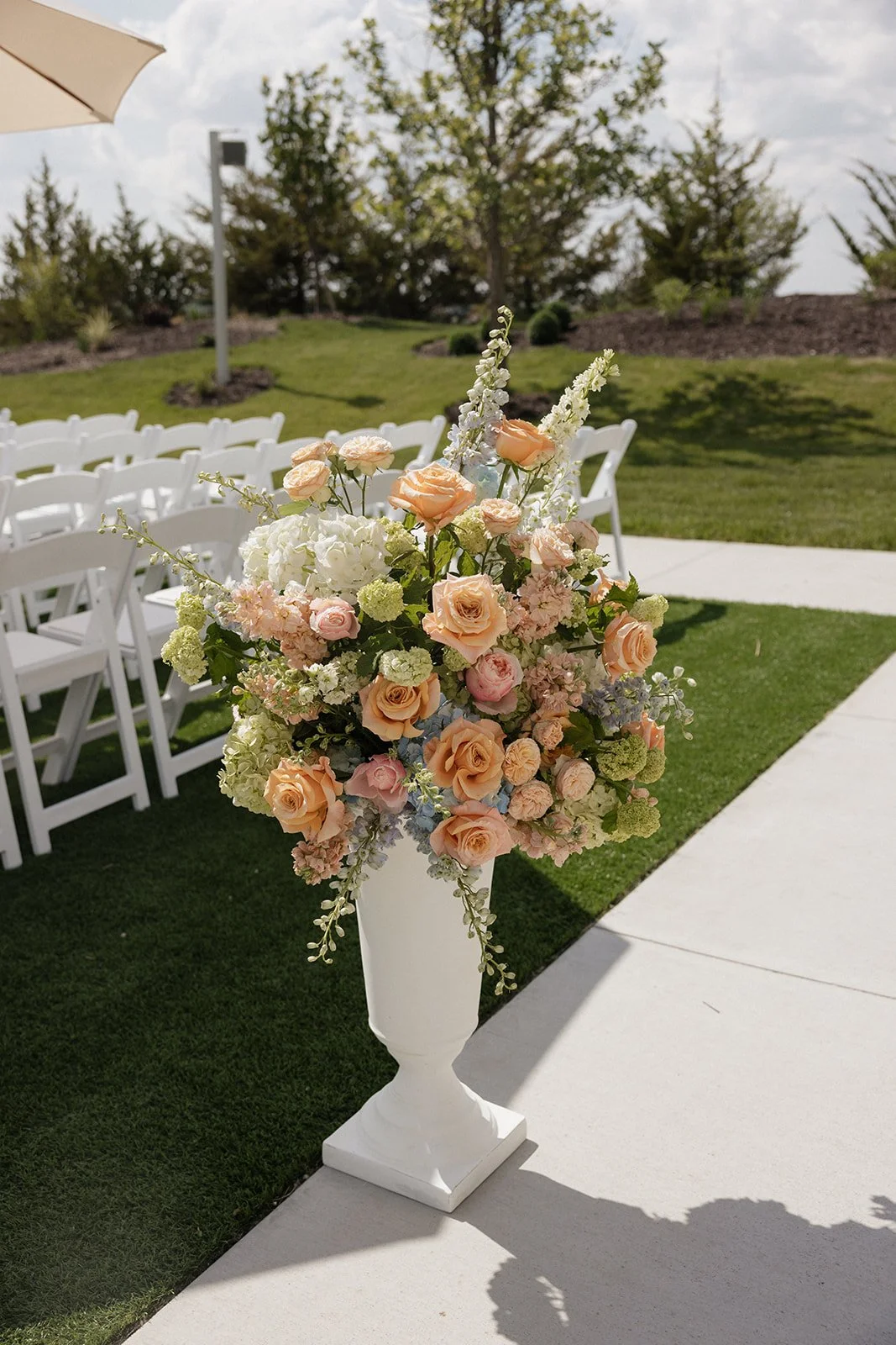 A large floral arrangement with peach and pink roses, white hydrangeas, and other flowers in a tall white vase set outdoors on a pathway, with chairs and trees in the background, suggesting a wedding or outdoor event setup.