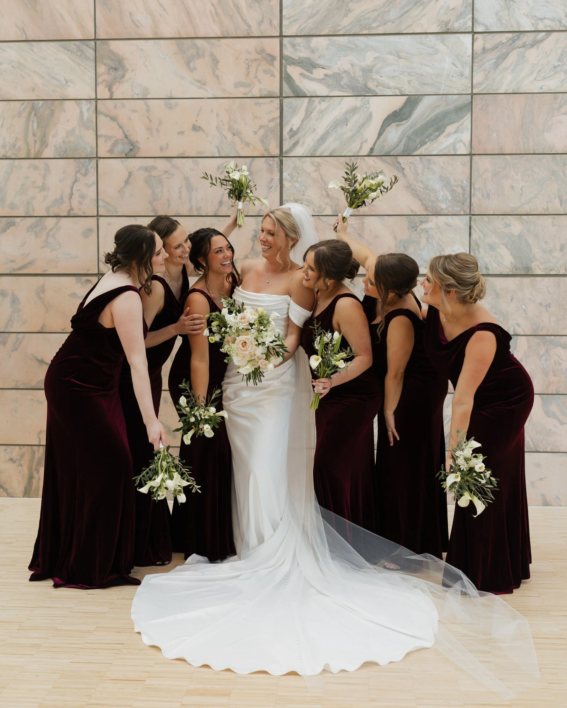Bride in a white wedding dress surrounded by her bridesmaids in dark burgundy dresses, all holding bouquets, standing against a marble wall.