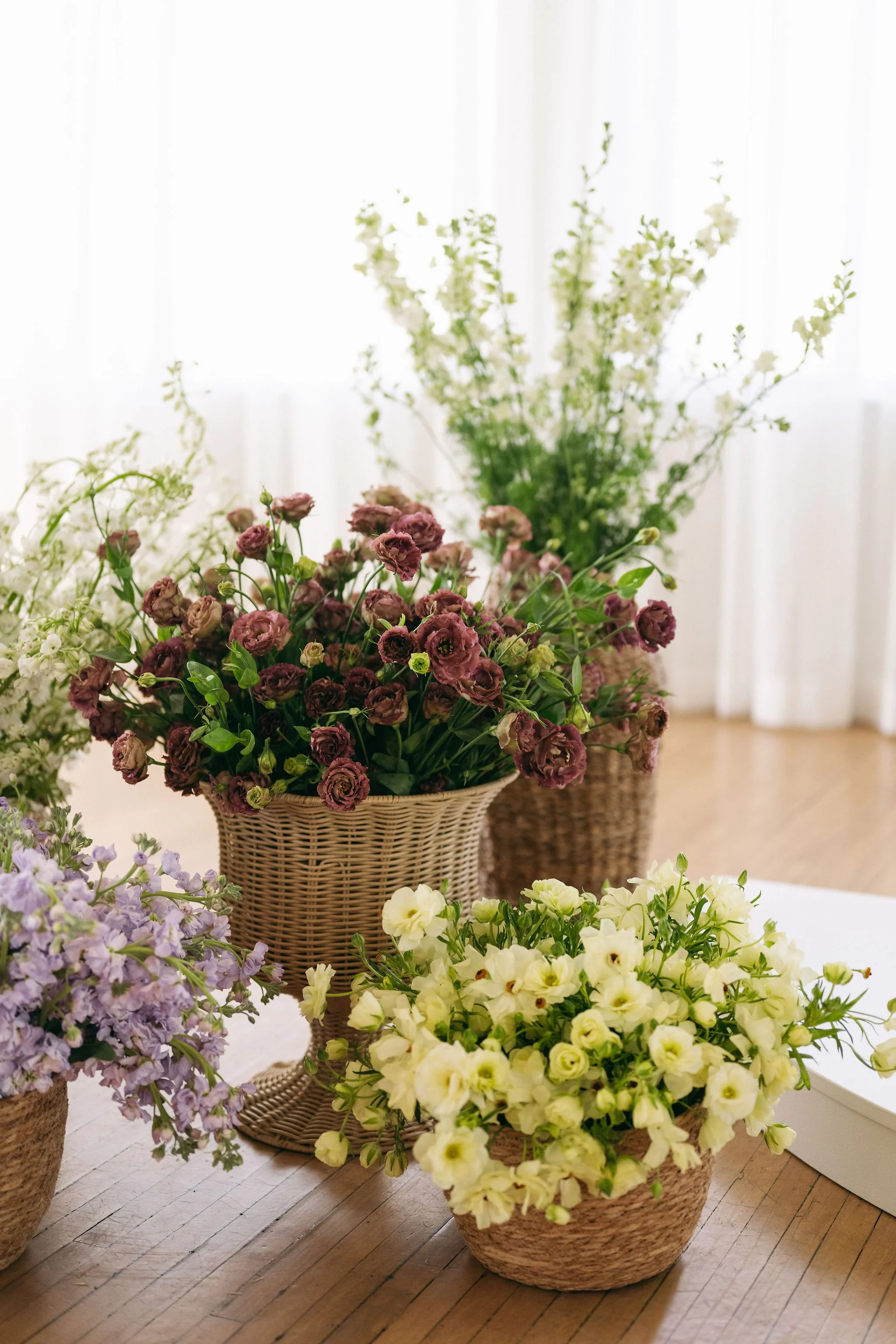 Several flower arrangements in woven baskets and planters on a wooden table, with a blurred bright window in background.