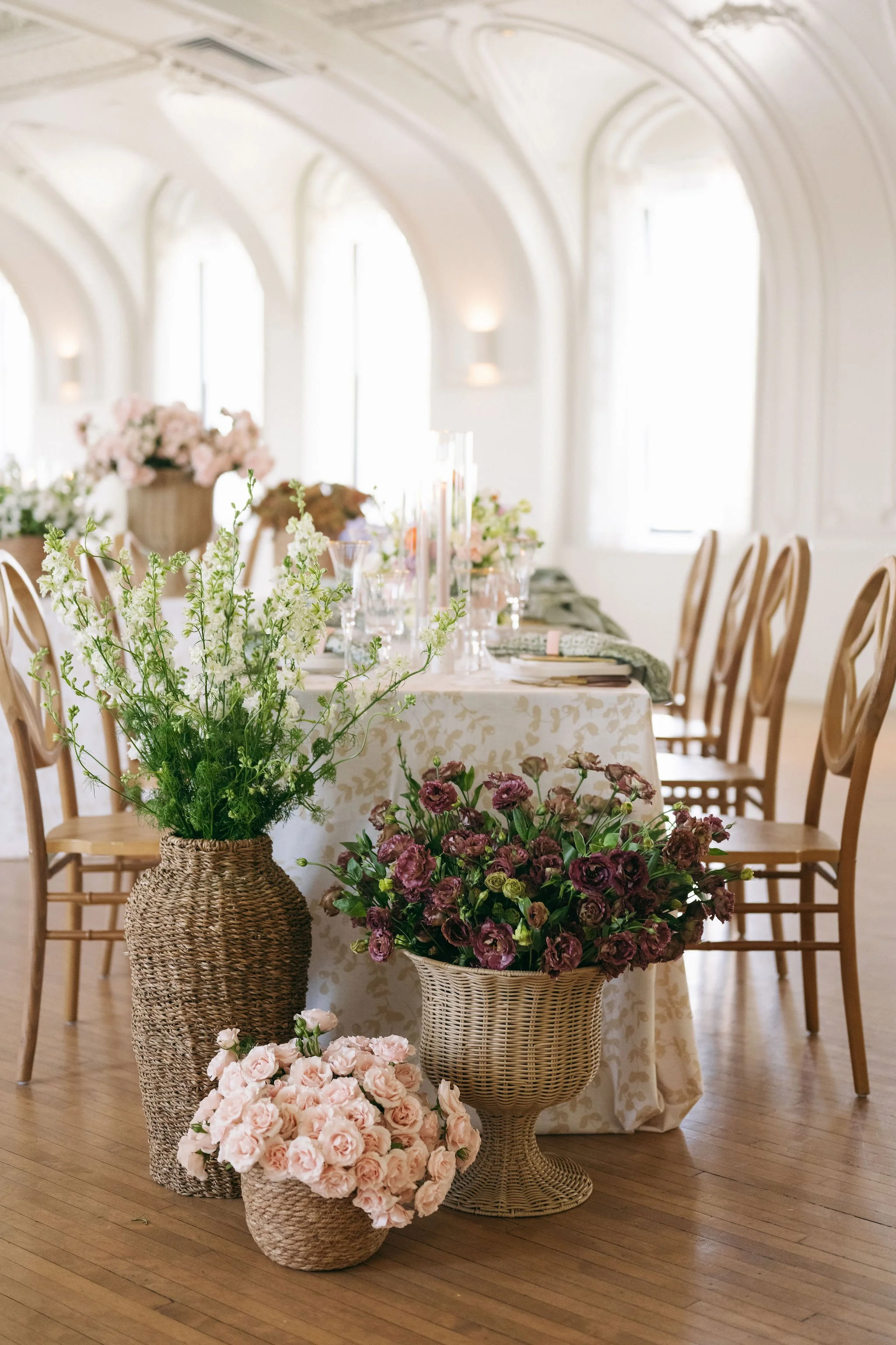 A decorated event space with a long table set for a celebration, adorned with floral arrangements in woven baskets and vases, including white, pink, and purple flowers. The room features high arched ceilings, large windows letting in natural light, a