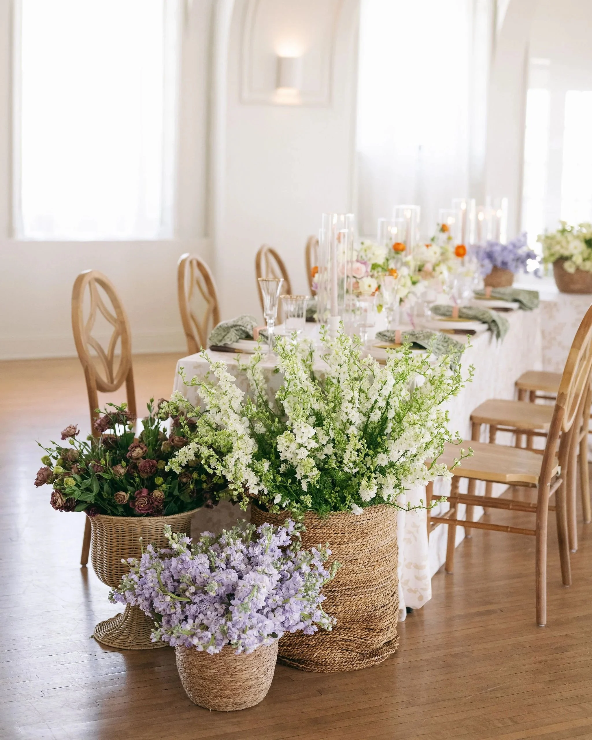 Elegant banquet table decorated with floral arrangements including white, purple, and pink flowers in wicker and woven baskets, set up with glassware and napkins in a bright, airy room.