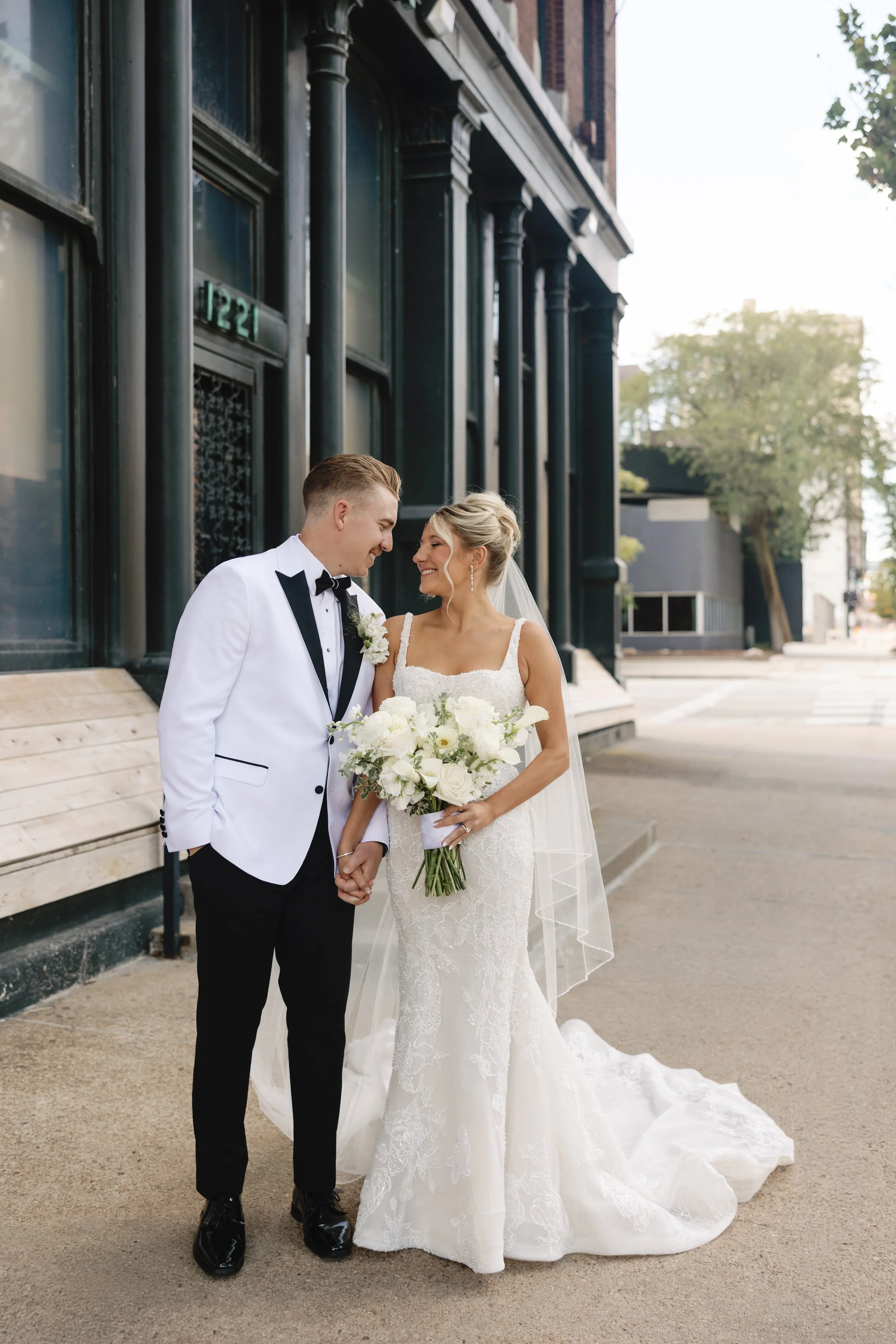A newlywed couple in wedding attire, holding hands and smiling at each other on a city sidewalk, with a dark building and trees in the background.