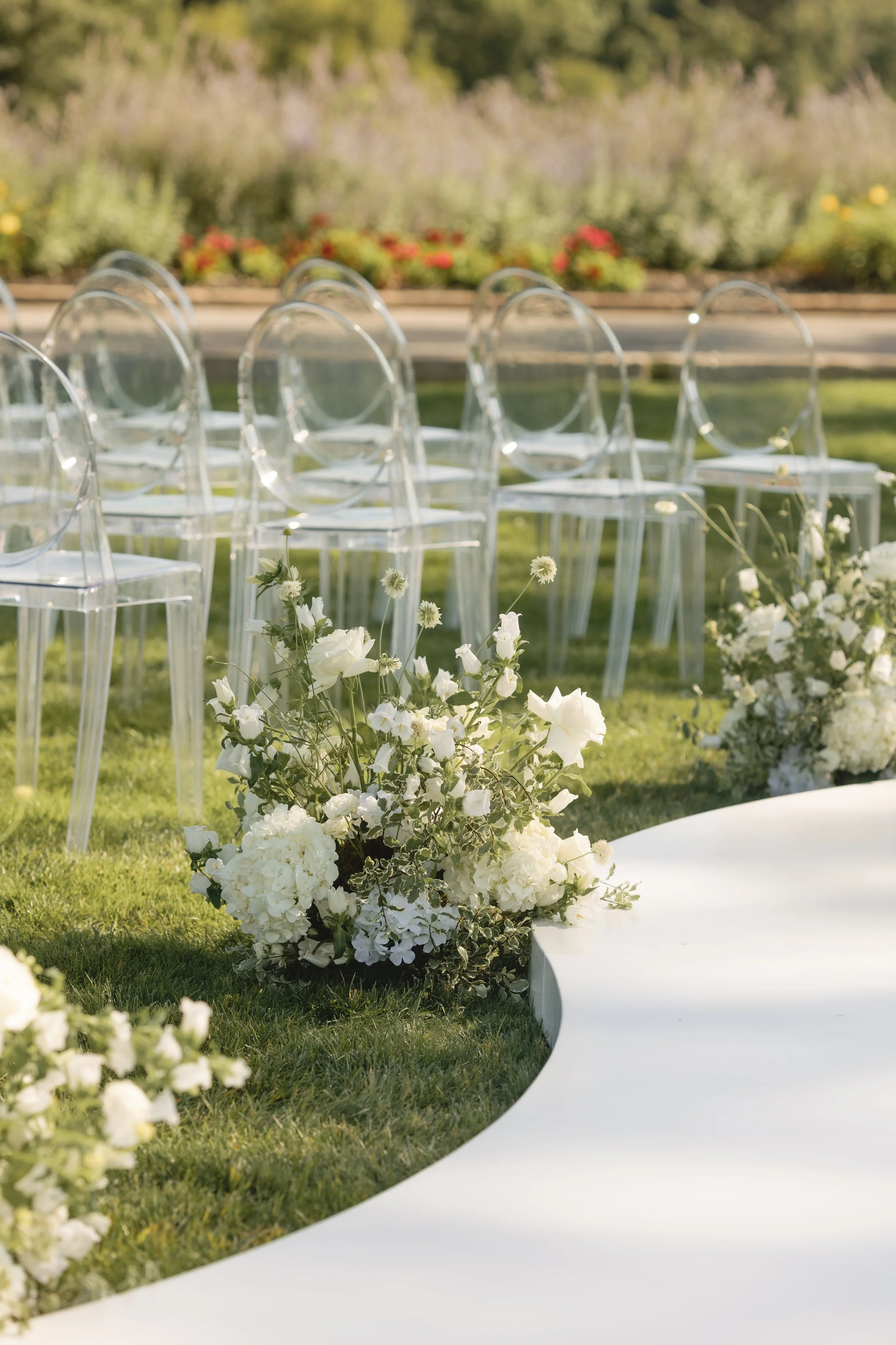 Outdoor wedding setup with clear chairs and white floral arrangements on green grass.