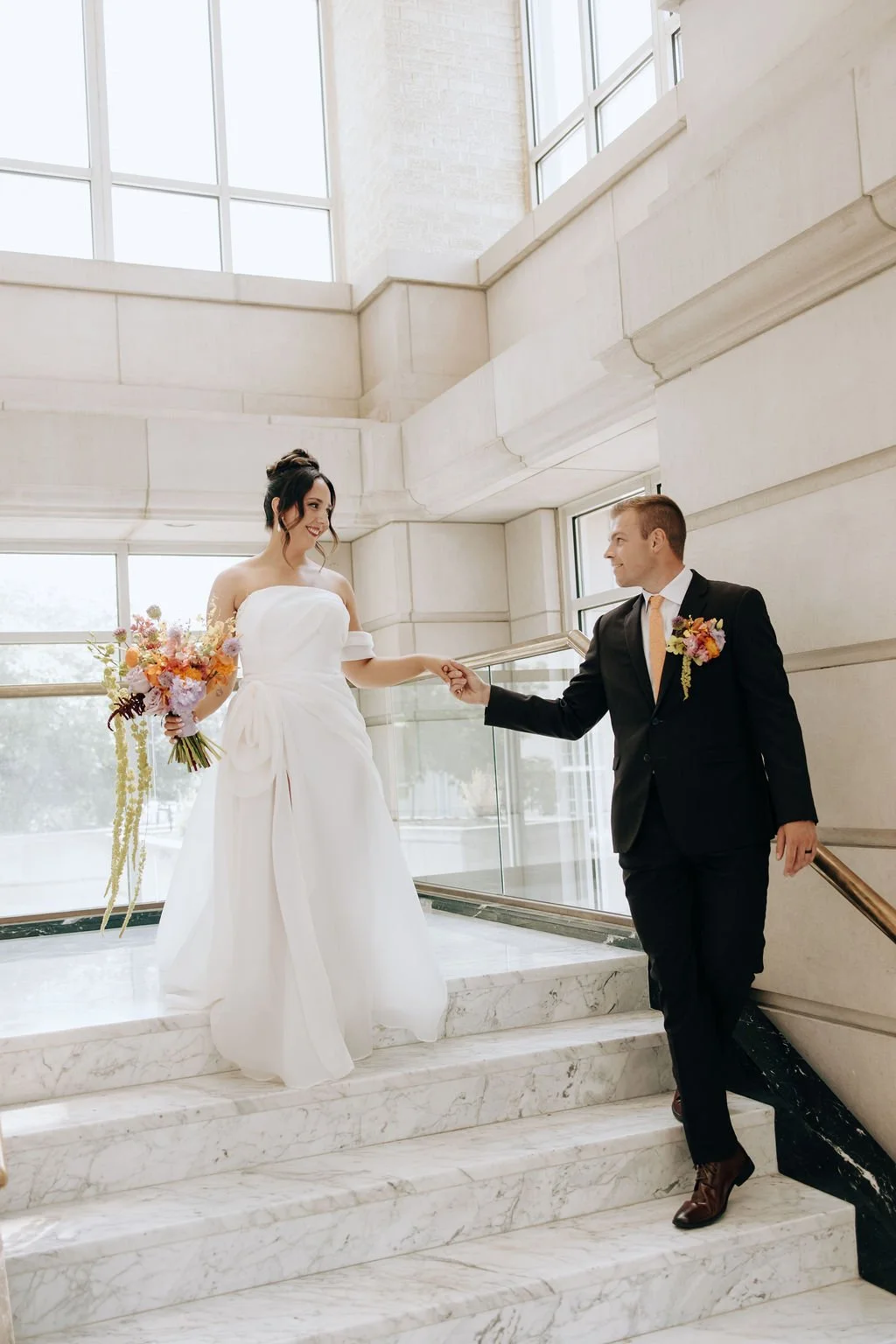 A bride in a white wedding dress holding a bouquet and a groom in a black suit with a boutonniere, holding her hand on a marble staircase inside a bright, modern building.