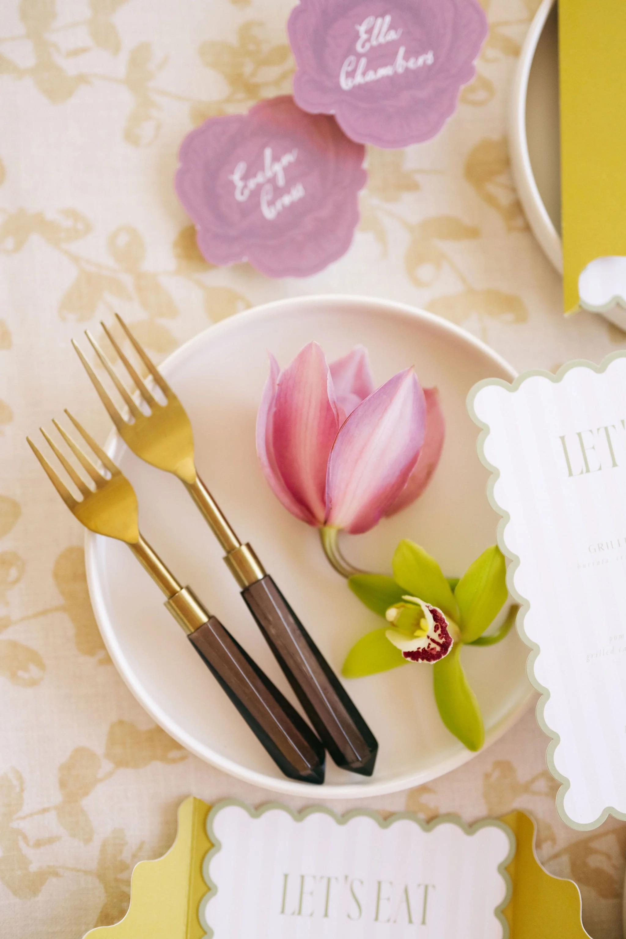 A white plate holding two gold forks with dark wood handles, a pink tulip, and a white orchid flower. Pink and purple flower-shaped name cards with 'Ella Chambers' are in the background.