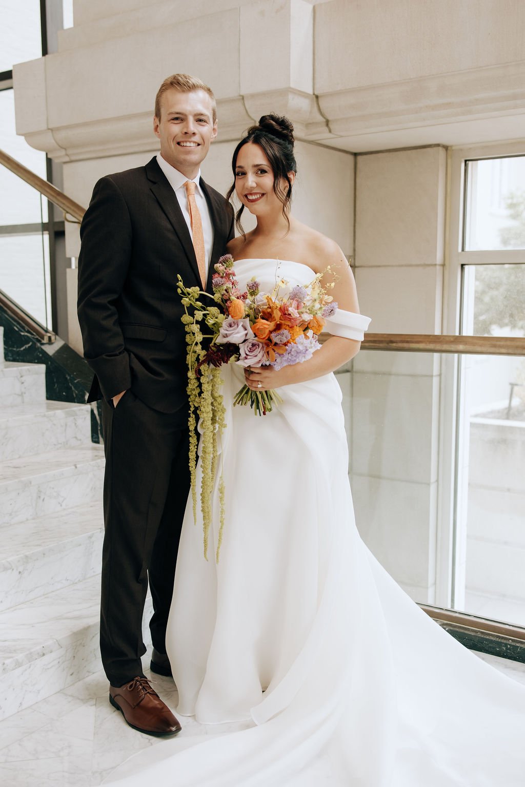 A newlywed couple posing on a marble staircase, the groom in a black suit and the bride in a white wedding dress holding a colorful bouquet, smiling indoors near large windows.