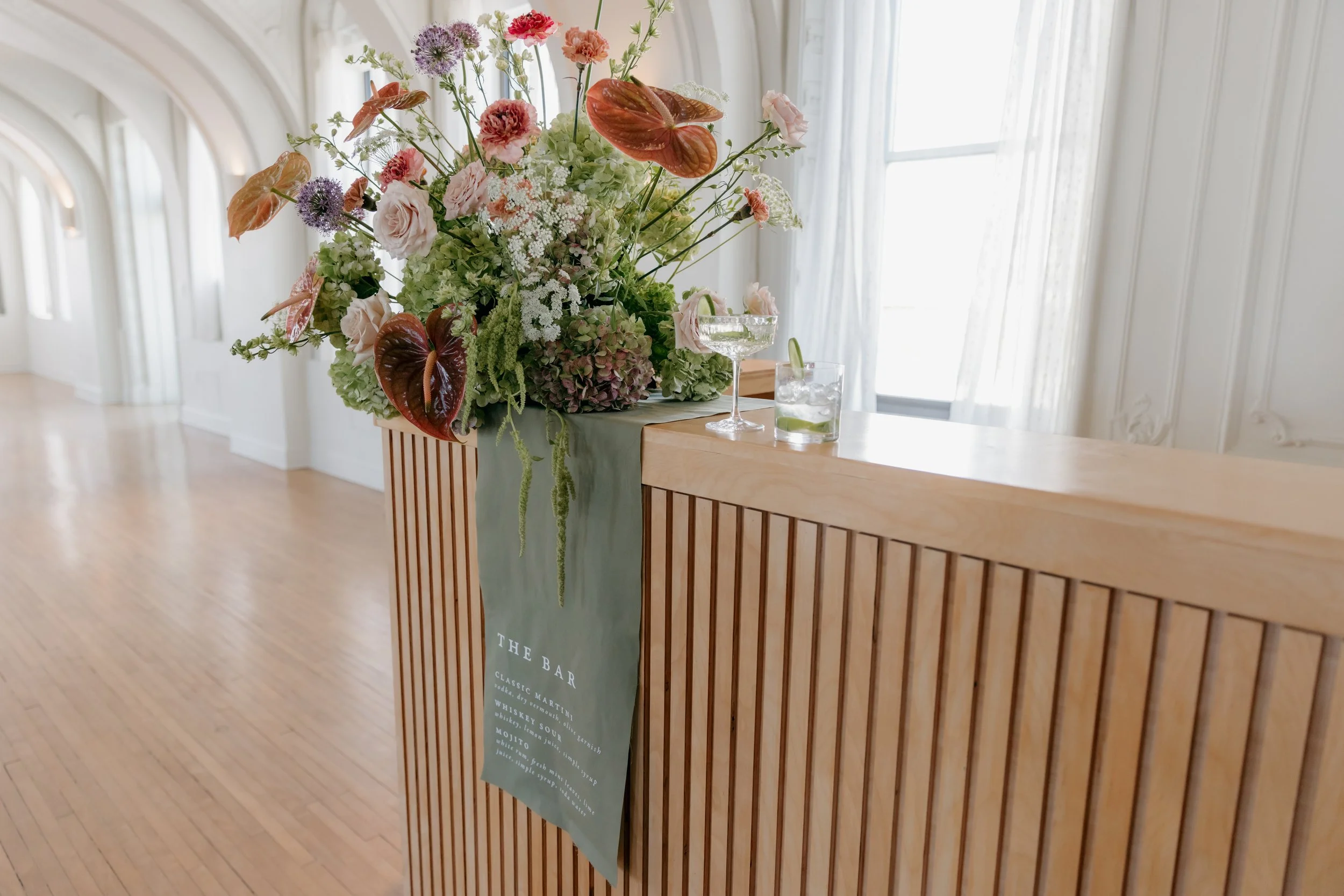 An elegant flower arrangement on a wooden bar counter in a bright, airy room with white walls and large windows.