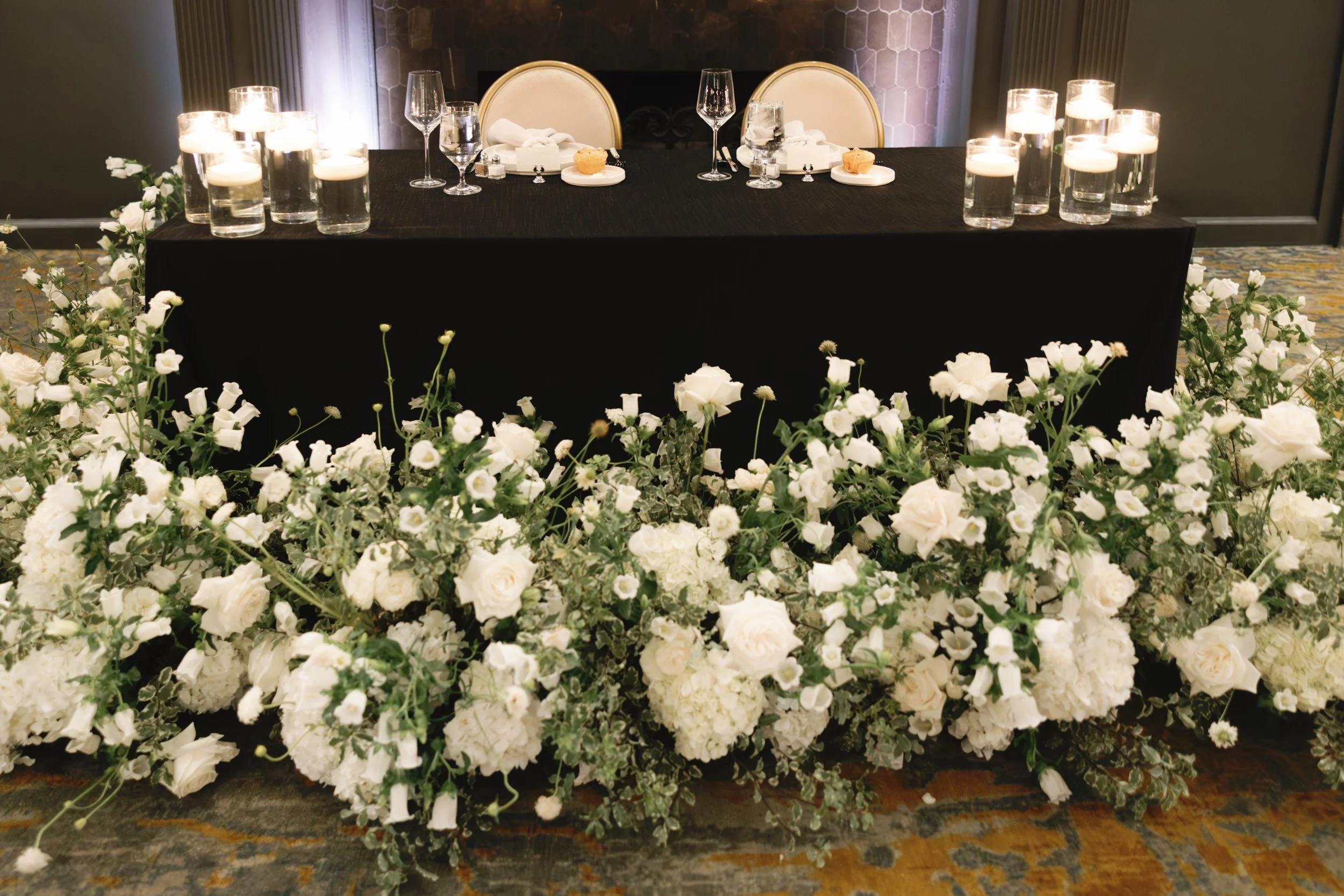 A wedding or event table adorned with tall candles and glassware, with a backdrop of a fireplace or wall, and decorated with a large arrangement of white flowers in front.