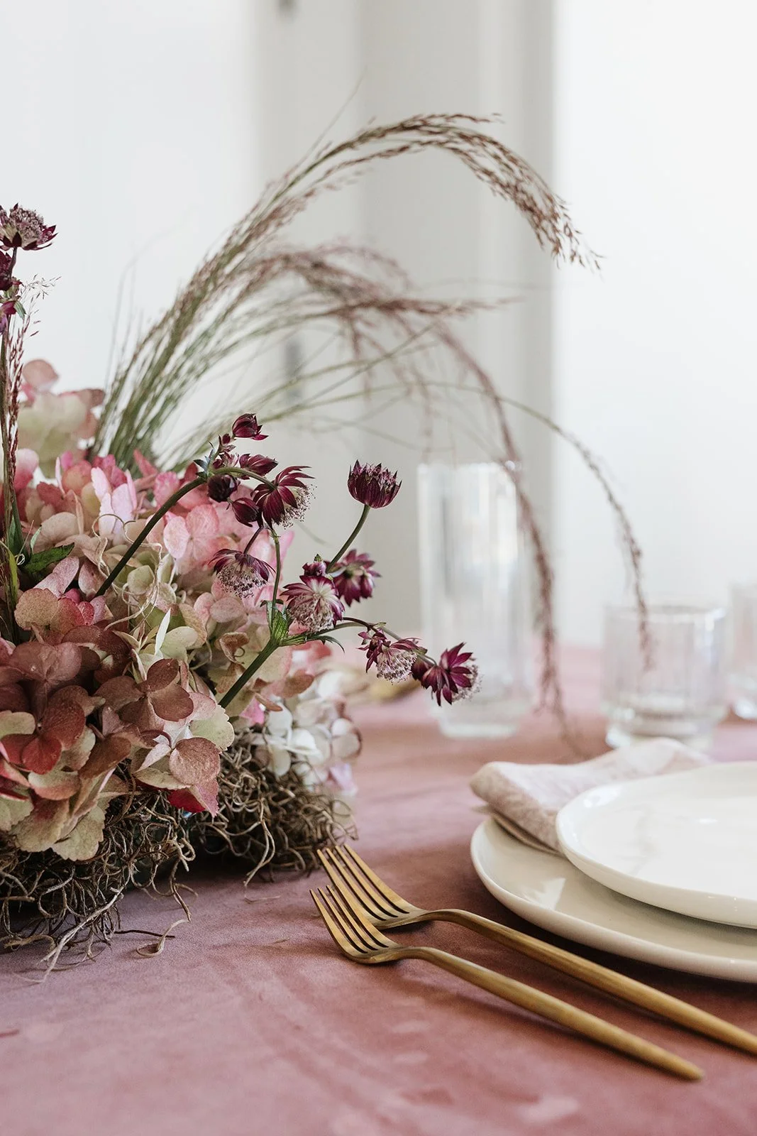 A close-up of a table setting with gold forks, white plates, a beige cloth napkin, and a floral centerpiece with pink and purple flowers and tall grasses in clear glass vases on a pink tablecloth.