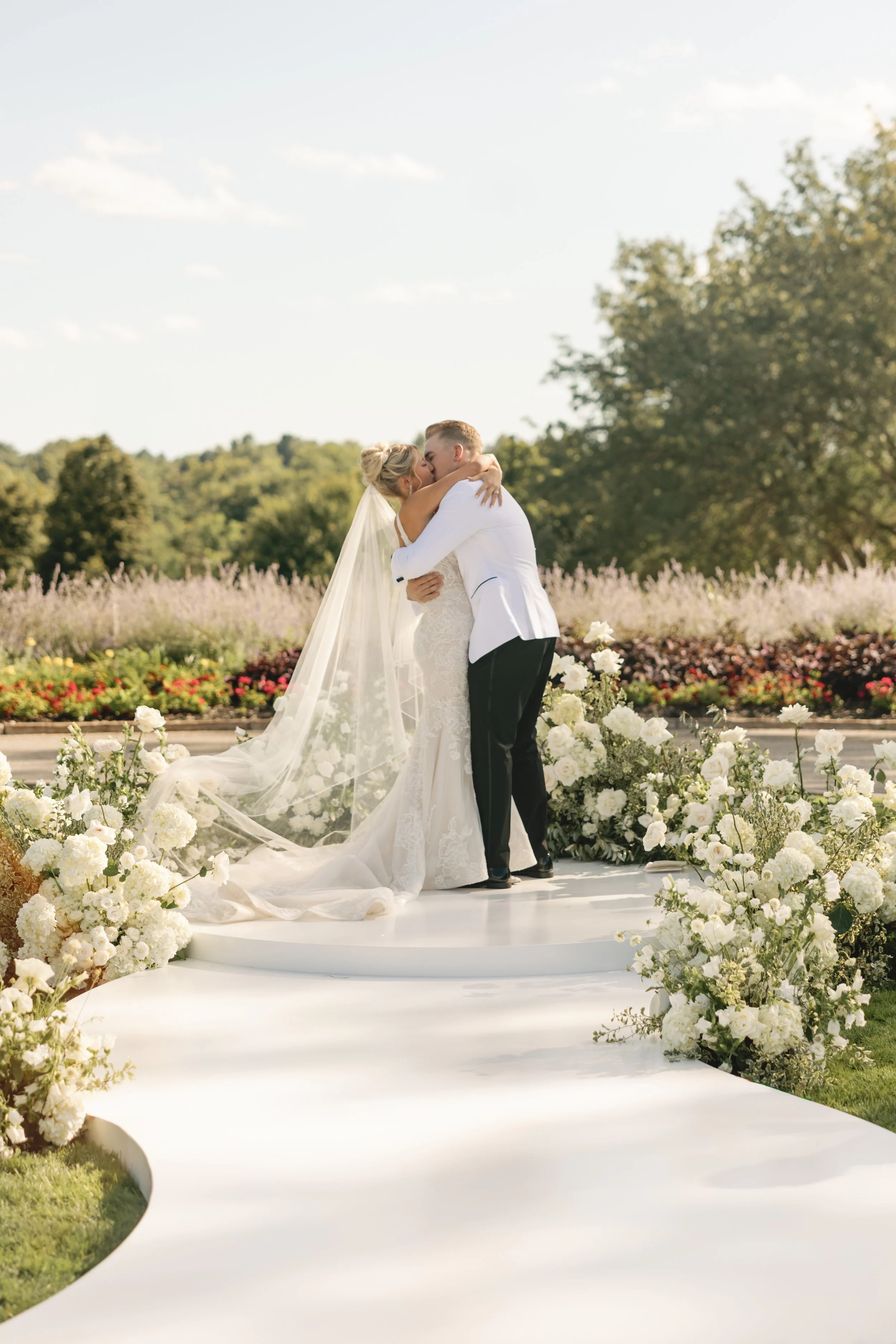 A bride and groom share a kiss on a wedding altar decorated with white flowers outdoors on a sunny day.