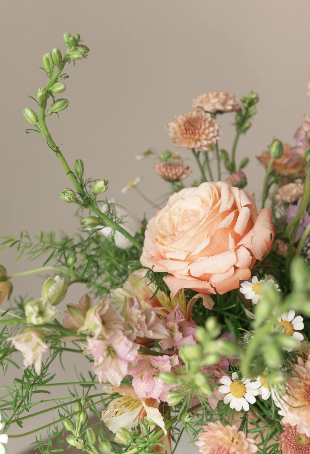 Close-up of a bouquet of mixed flowers including a large peach-colored rose, pink and white daisies, and other small pink and white flowers with green foliage.
