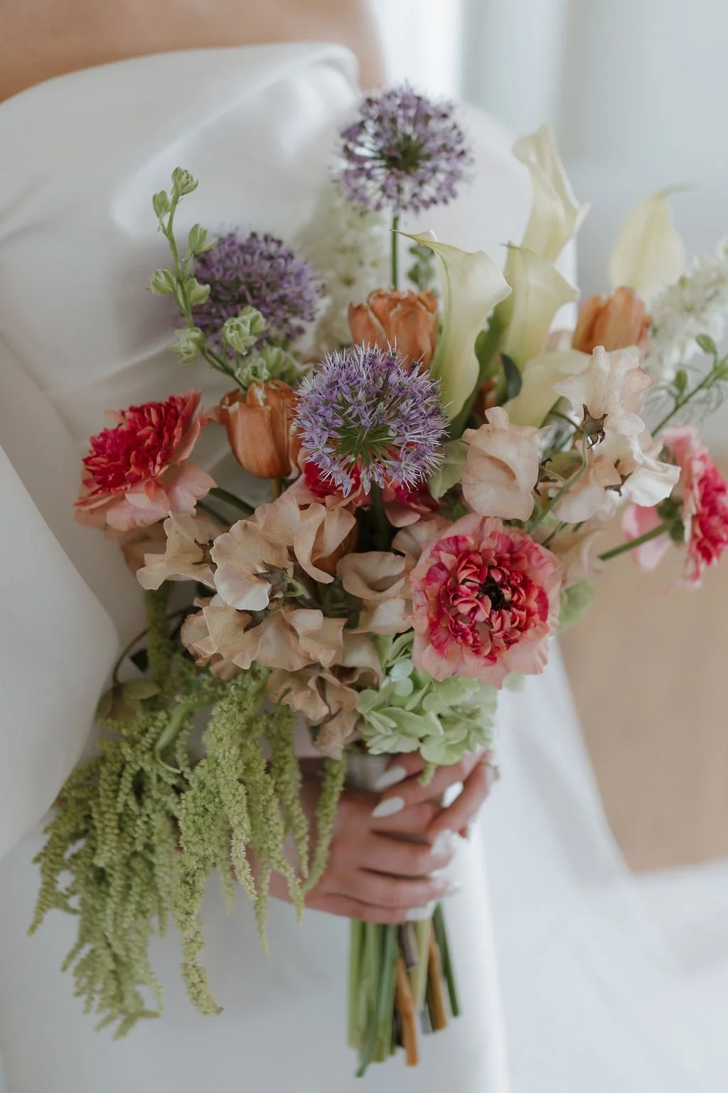 Close-up of a vibrant mixed flower bouquet held by a person in a white dress, with a soft-focus background.